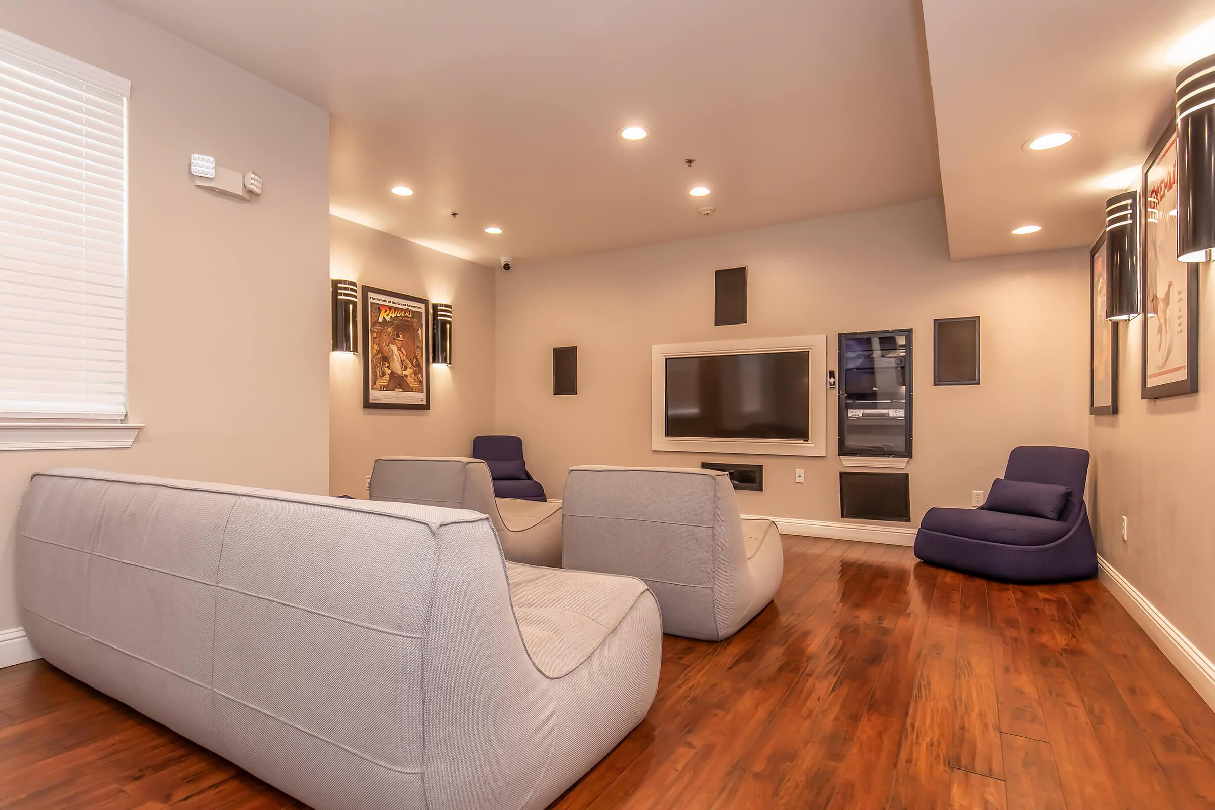A modern living room featuring two gray sofas and a comfortable blue chair. The walls are painted in light neutral tones with ambient lighting, and there are framed posters on the walls. A wall-mounted television is positioned centrally, creating a cozy entertainment space with hardwood flooring.