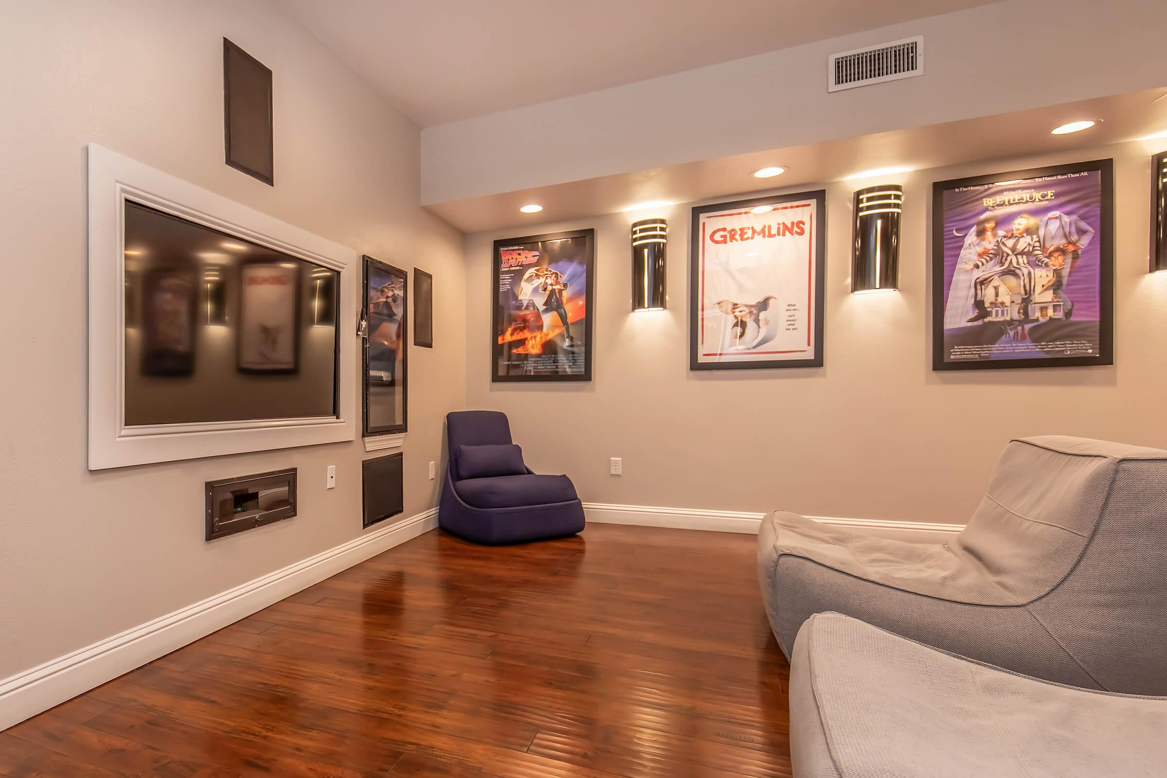 A cozy home theater featuring a large flat-screen TV mounted on the wall, surrounded by framed movie posters for "Gremlins" and other films. The room has hardwood flooring, with a modern gray sectional sofa and a blue lounge chair, all under soft ambient lighting.