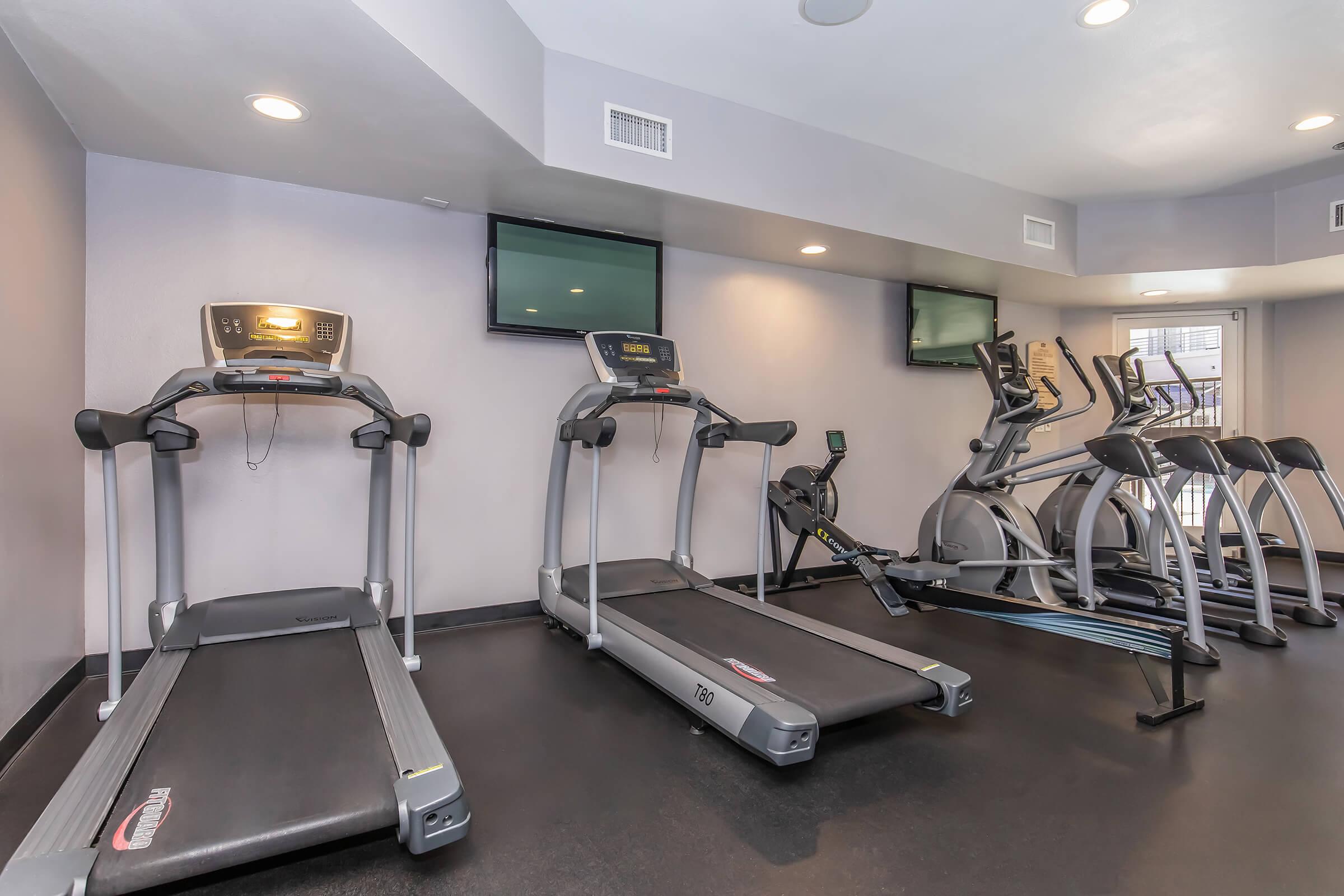 A modern fitness gym with several exercise equipment pieces, including two treadmills and an elliptical machine. The space features light-colored walls, a black floor, and multiple flat-screen TVs mounted on the walls for entertainment. Bright lighting enhances the workout environment.