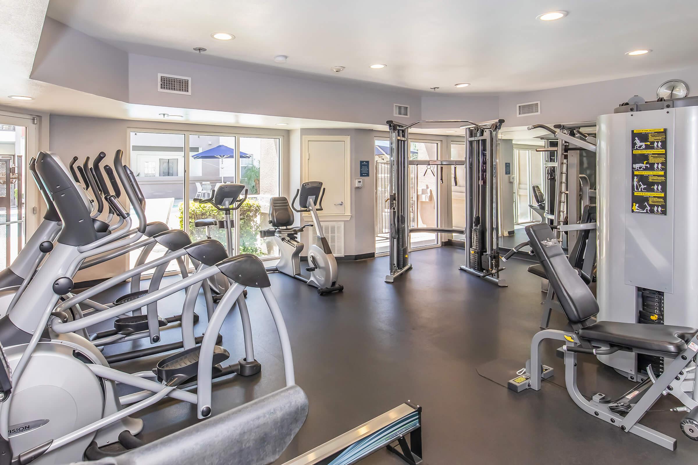 A modern fitness center featuring various exercise equipment, including stationary bikes, weight machines, and a spacious layout with large windows letting in natural light. The room has a clean and inviting atmosphere, suitable for workouts.
