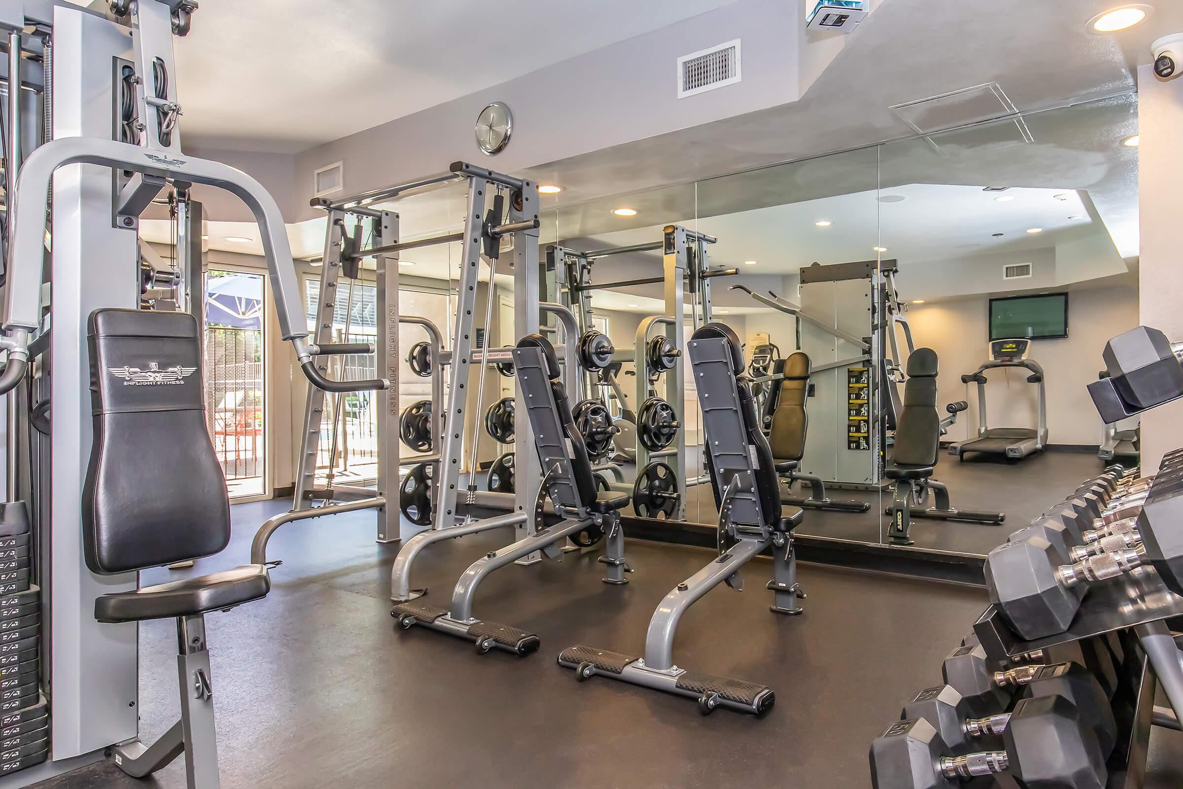 A modern gym featuring various workout equipment, including weight machines and free weights. The space is well-lit with large mirrors on one wall, showcasing the equipment. There are also treadmills and other fitness machines visible in the background, creating an inviting atmosphere for exercise.