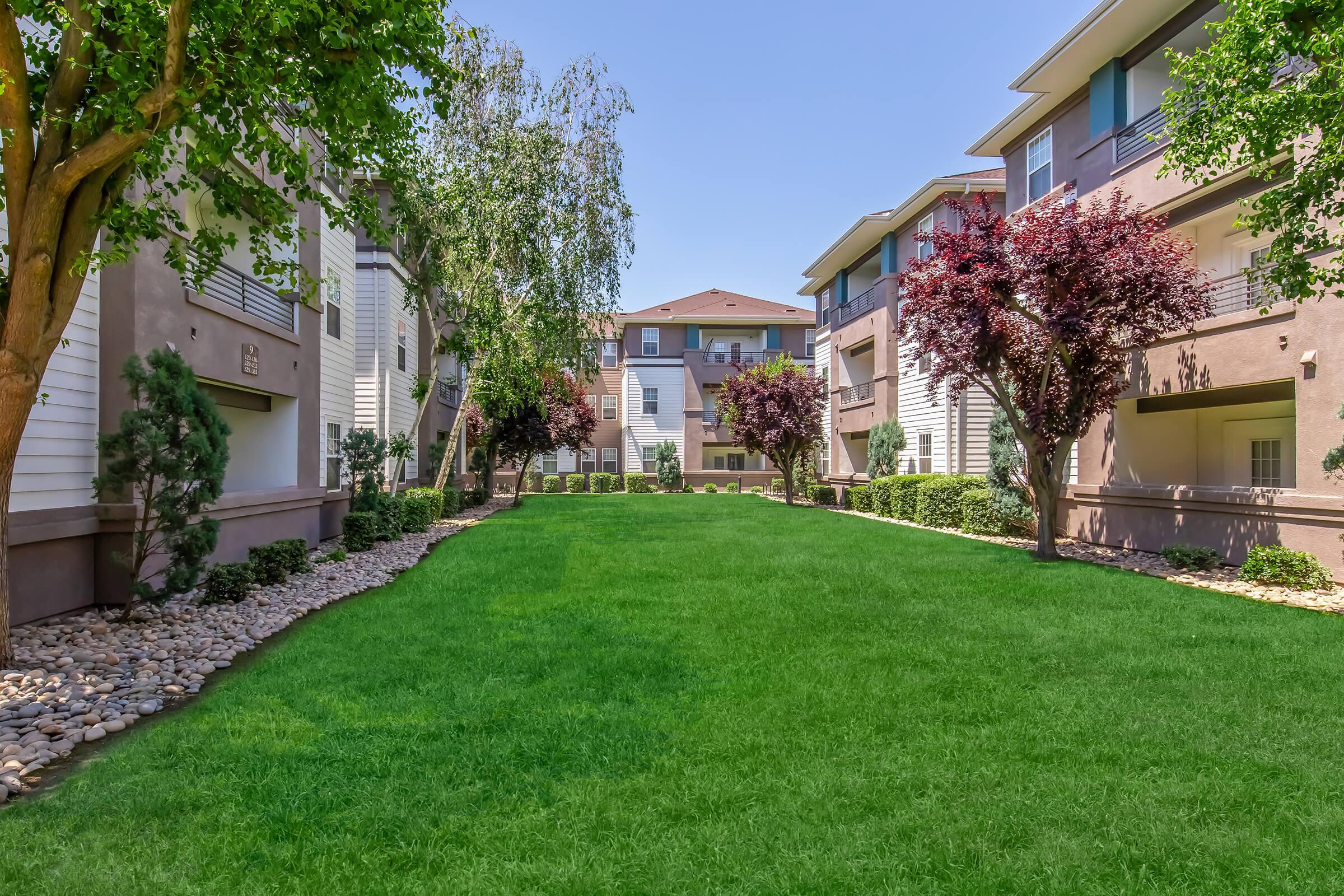A well-maintained green lawn surrounded by modern apartment buildings. The landscape features neatly trimmed bushes and trees, and a clear blue sky overhead. The area is designed for relaxation and enjoyment, providing a serene atmosphere.