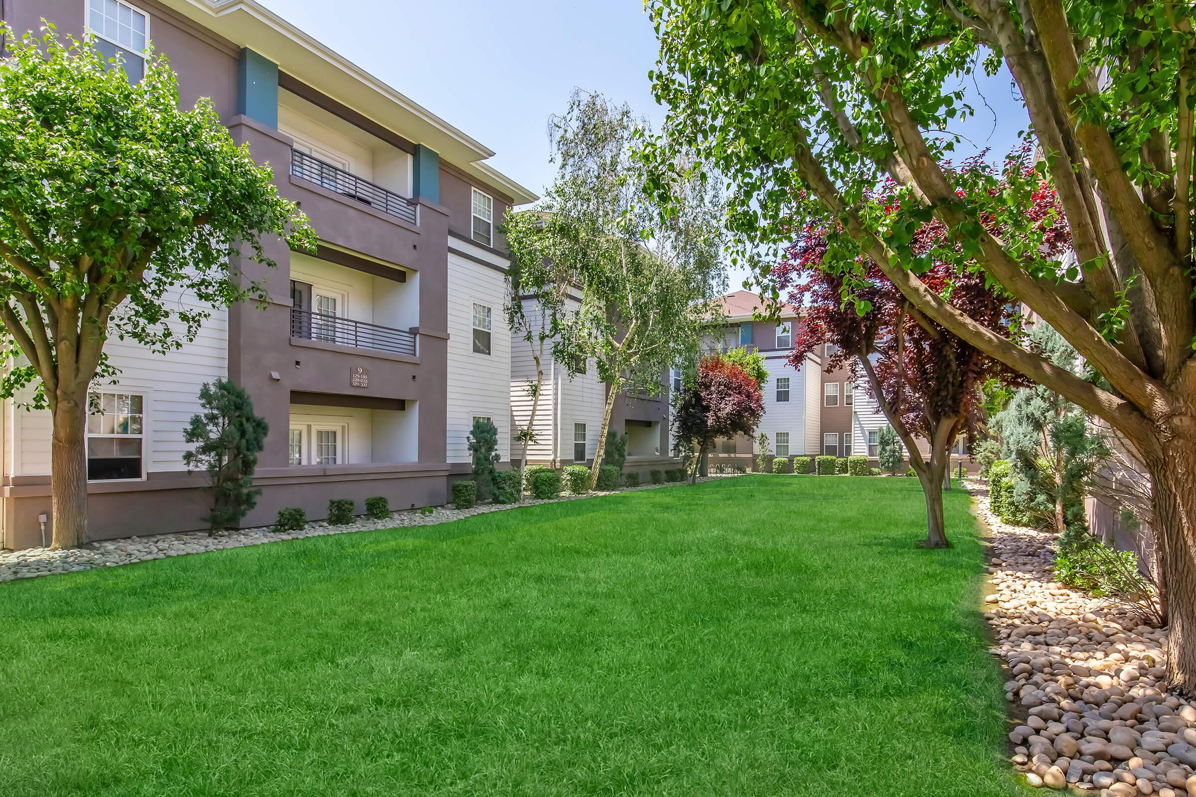 A well-maintained apartment complex surrounded by lush green grass and landscaped trees. The scene features two buildings with varied architectural designs, creating a serene outdoor environment. Pebbles line the pathway, adding a decorative touch to the landscaped area.