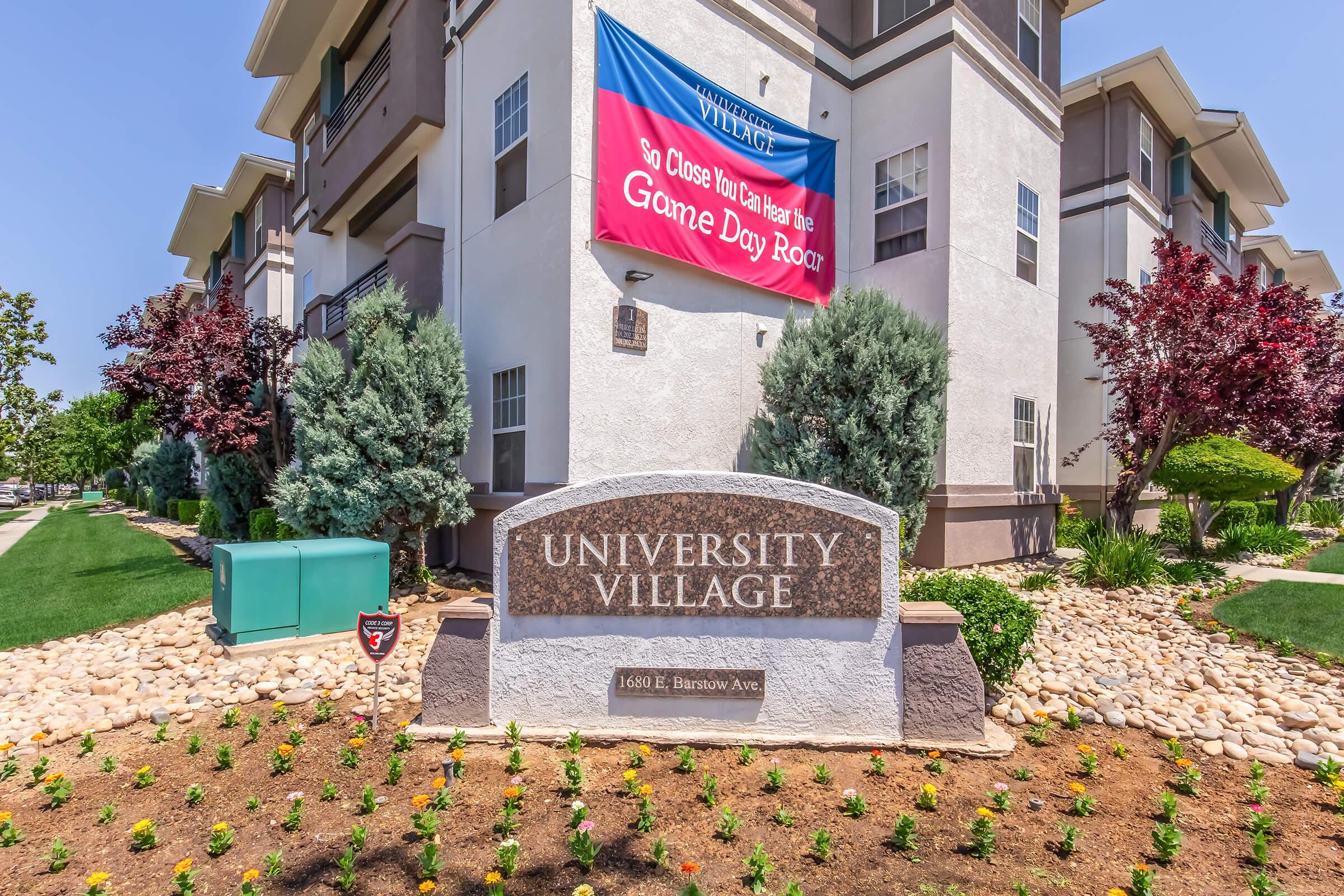 Sign for University Village located at 1600 E Barrow Ave, featuring a colorful banner that reads "So Close You Can Hear the Game Day Roar". The area is landscaped with shrubs, flowers, and includes nearby buildings typical of an apartment complex.