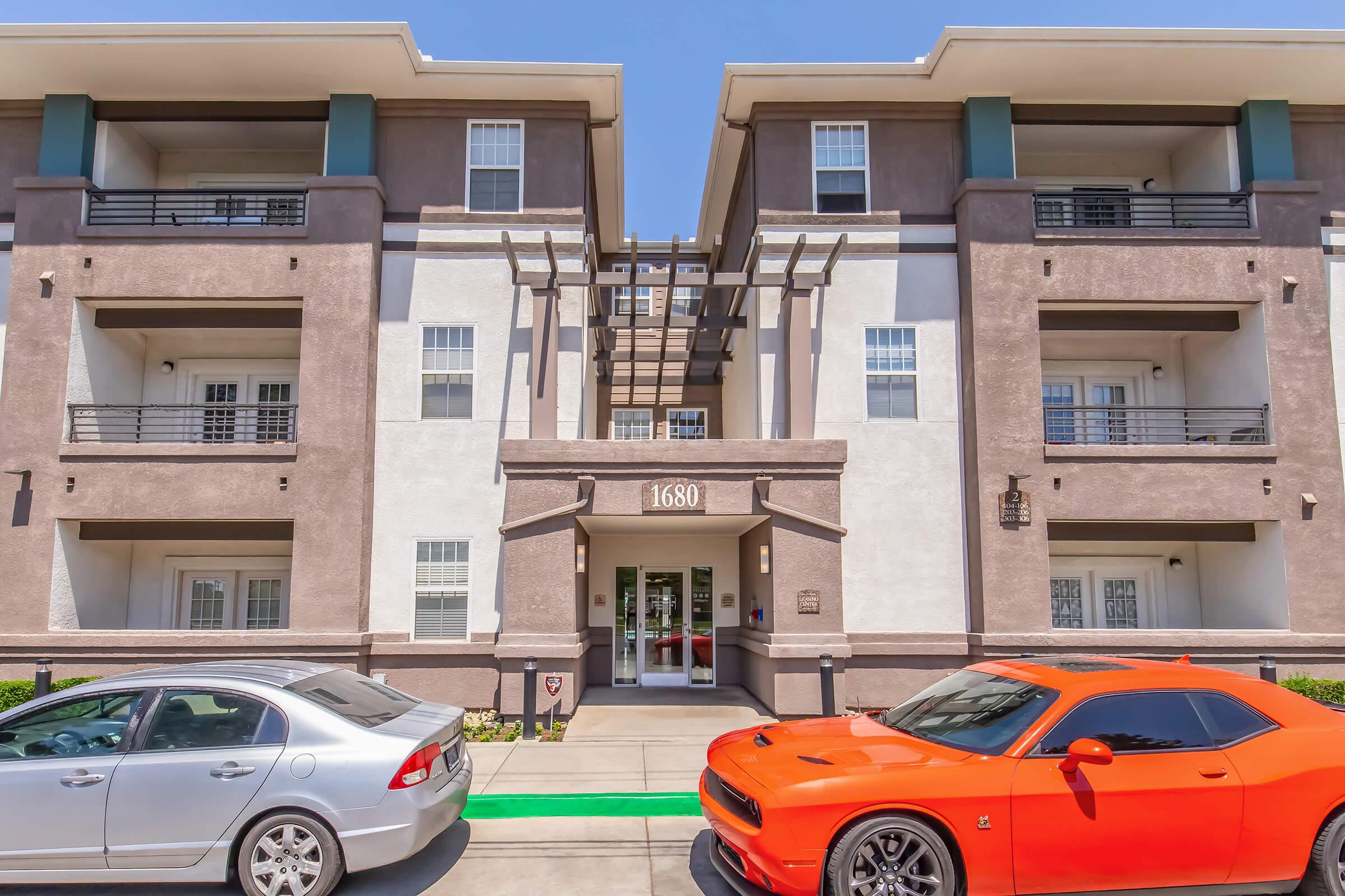 A modern apartment building with three stories, featuring a mix of brown and white exterior colors. The entrance is centered, flanked by balconies on the upper floors. Two parked cars, a silver sedan and an orange sports car, are visible in front. Clear blue sky overhead.