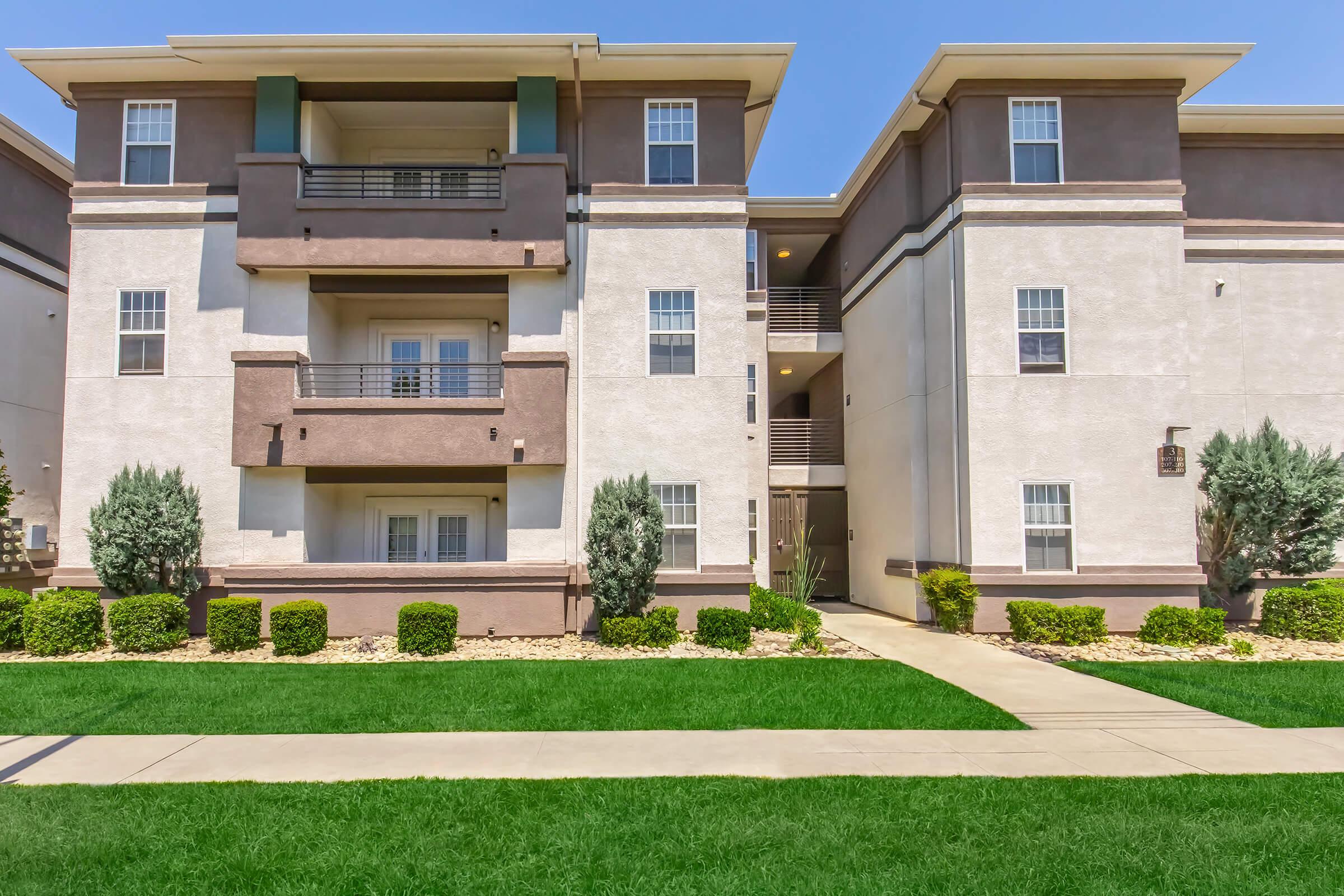 Three-story apartment building with a modern design, featuring a combination of light and dark exterior walls. The ground level has a pathway leading to the entrance, surrounded by well-maintained grass and shrubs. There are balconies for each unit on the upper floors, with windows reflecting sunlight.