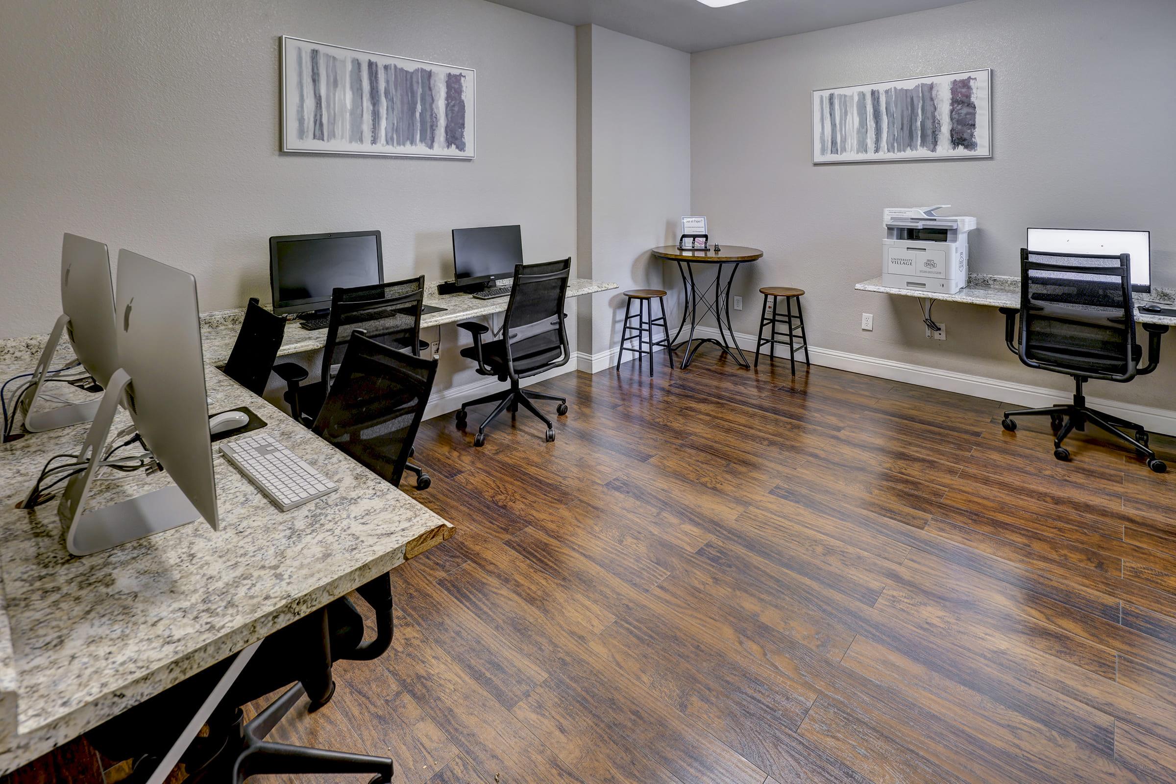 A modern office space featuring multiple workstations with desktop computers on granite-top desks, ergonomic chairs, and a printer. The floor is hardwood, and the walls are painted in neutral tones with abstract artwork hanging above the desks. Two stools are positioned near a standing workspace.