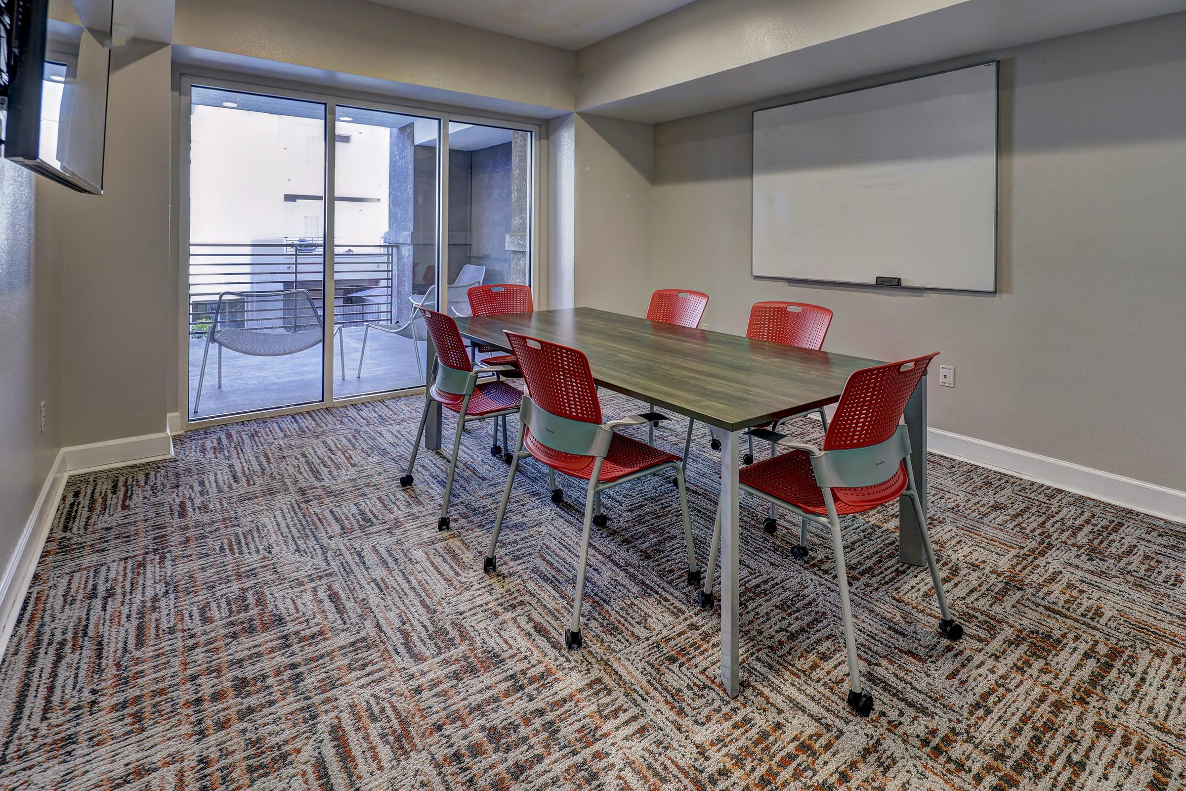 A conference room featuring a rectangular table surrounded by six red chairs. There is a whiteboard on one wall and large sliding glass doors leading to an outdoor area. The floor is covered with a patterned carpet, and a television is mounted on one wall. Natural light enters from the outside.