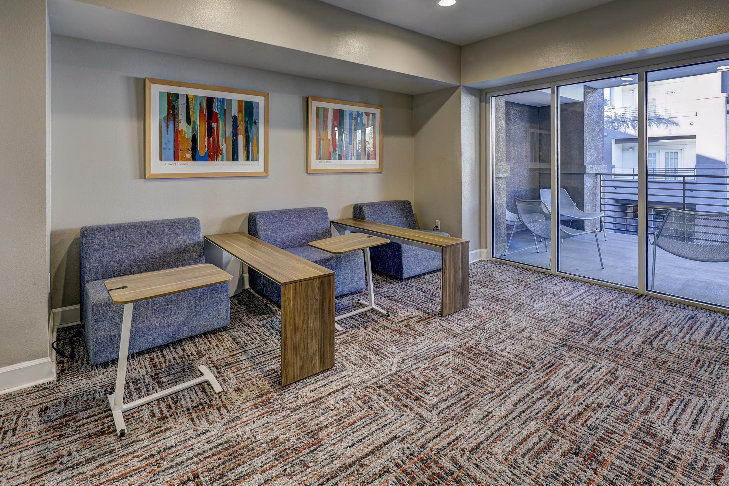 A modern lounge area featuring two blue chairs with small wooden tables between them. Framed colorful abstract artwork hangs on the wall, and large windows lead to an outdoor balcony, allowing natural light to fill the room. The flooring is covered with a patterned carpet.