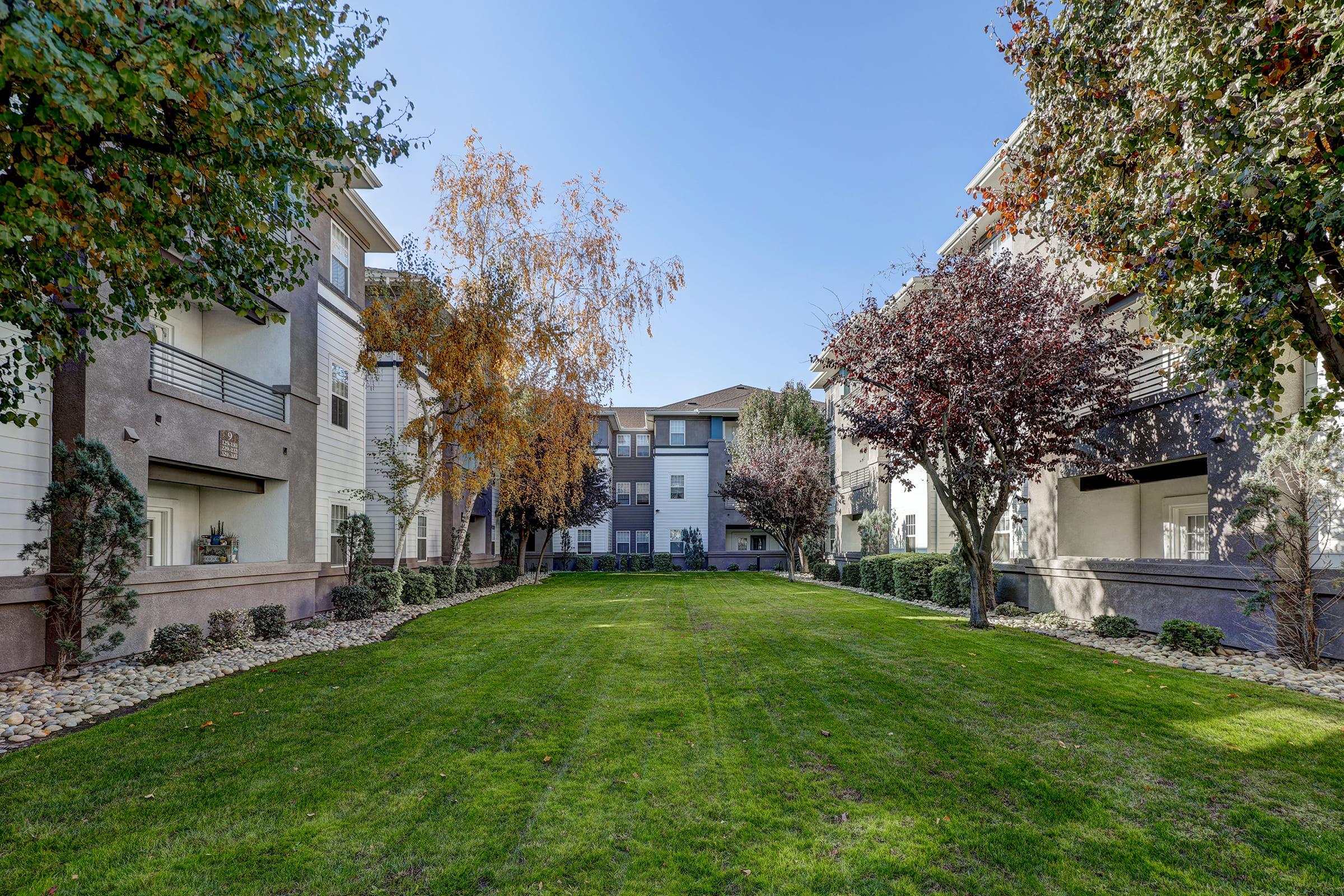 View of a landscaped courtyard surrounded by residential buildings. The green lawn is bordered by trees with autumn foliage, and flower beds create a serene atmosphere. Clear blue sky overhead, offering a bright and open environment.