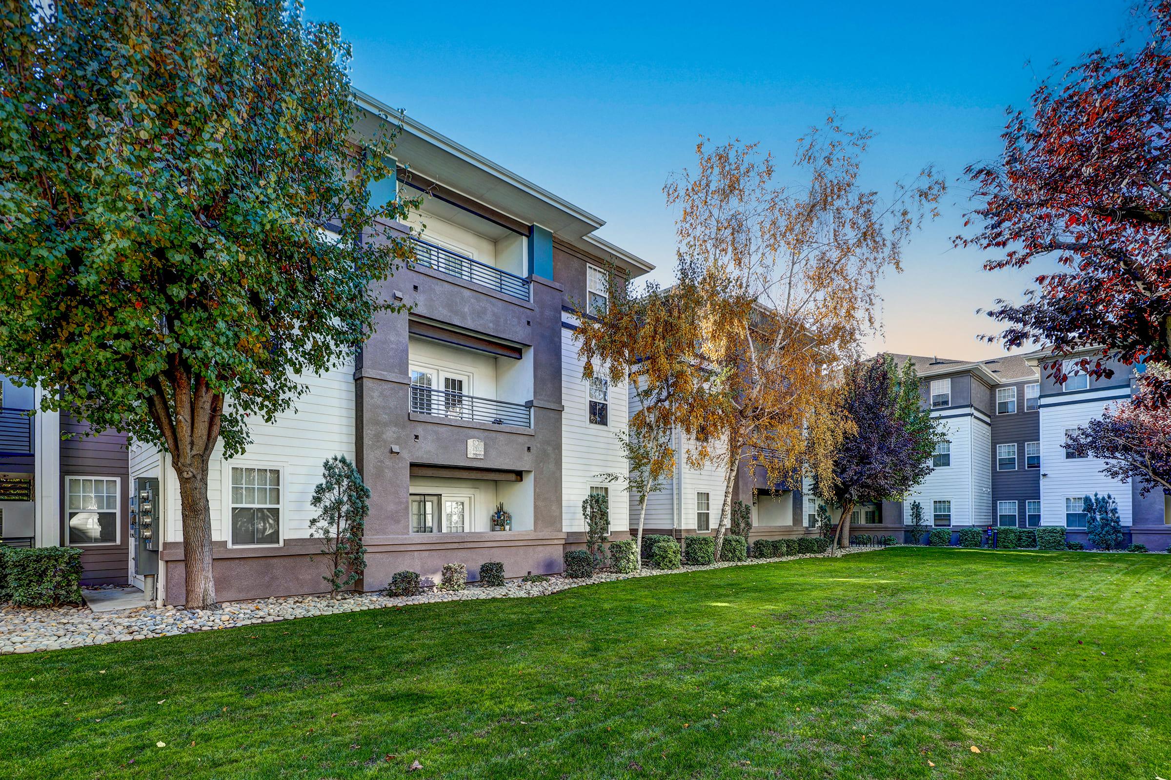 A well-maintained apartment community featuring modern architecture surrounded by lush green lawns. Trees with autumn foliage line the pathway, adding color to the setting. The building has multiple balconies and large windows, creating a welcoming atmosphere. The sky above is clear and blue, indicating a pleasant day.