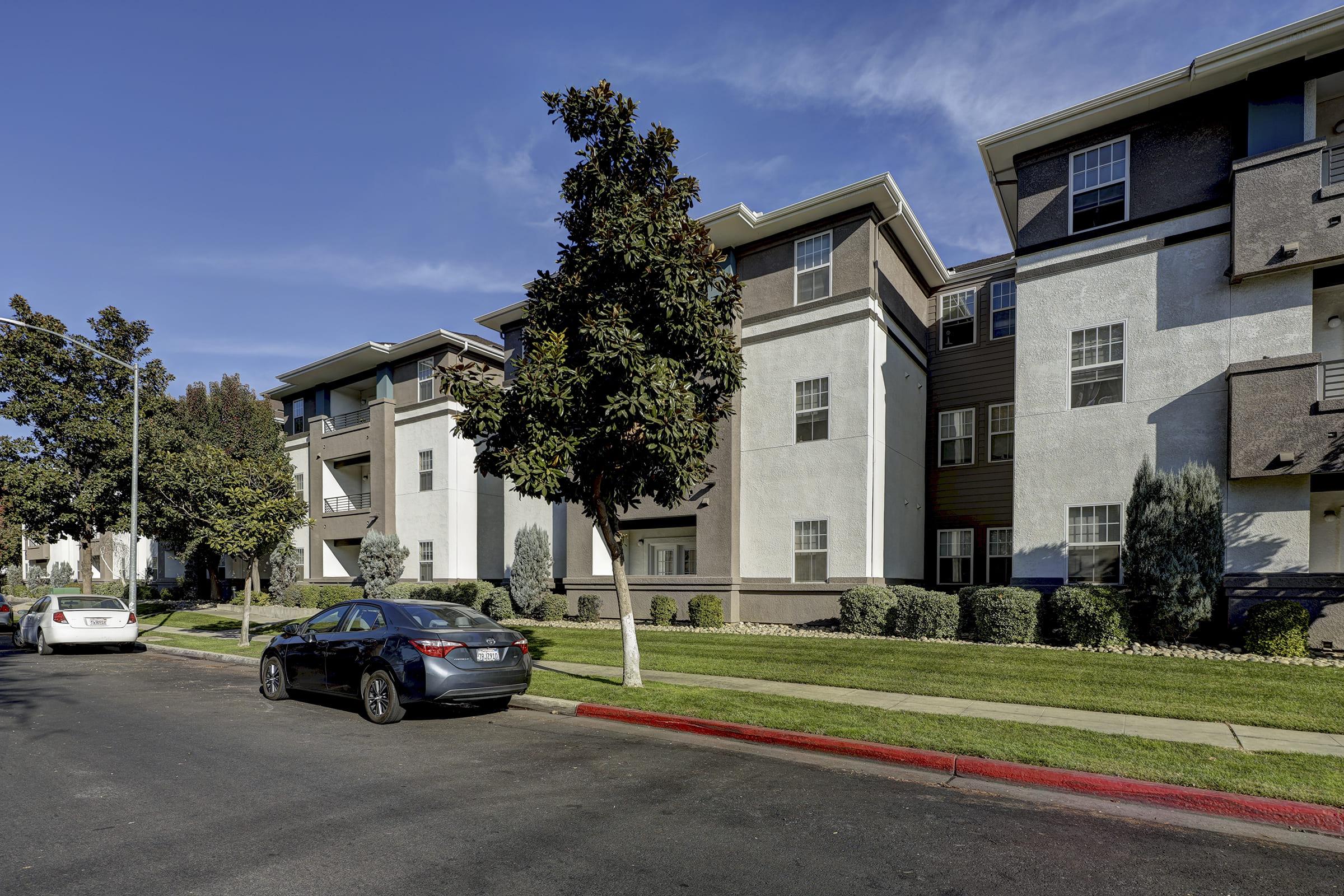 A view of a residential apartment community featuring three-story buildings with a combination of light and dark exterior colors. The scene includes well-maintained lawns and landscaping, with parked cars along the street and trees lining the sidewalk on a clear day.