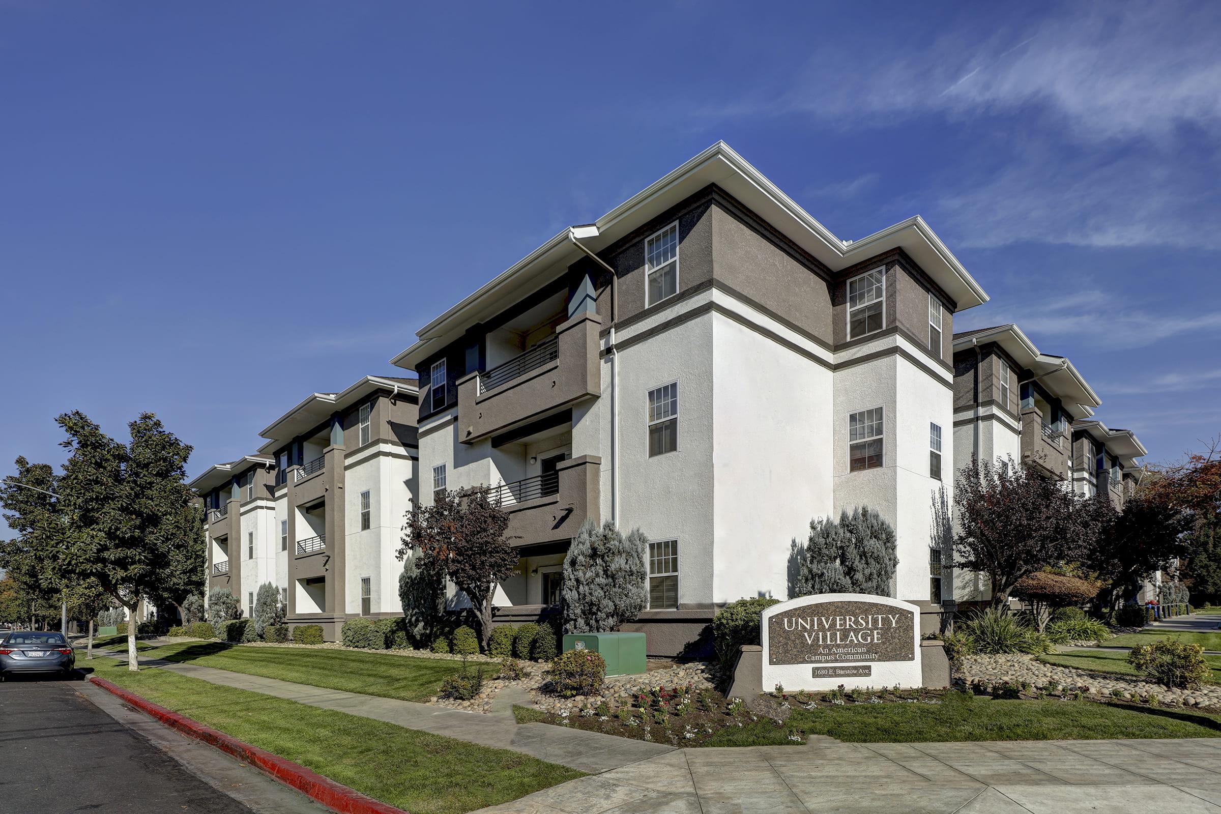 A view of the University Village apartment community, showcasing a modern, multi-story building with a welcoming entrance sign. The surrounding area includes landscaped greenery and a clear blue sky overhead. Parking spaces are visible on the side of the complex.