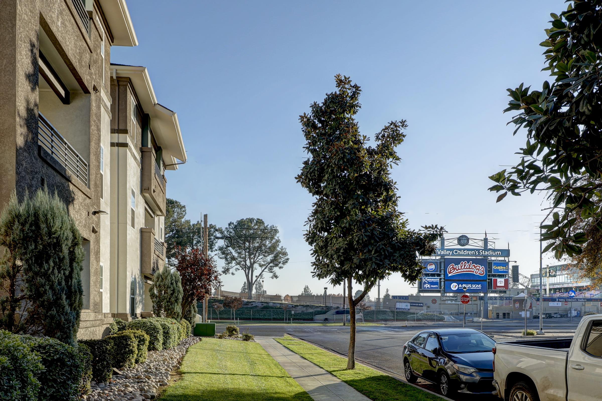 A street view showcasing a sunny day, featuring an apartment building on the left, well-maintained landscaping, and a large billboard advertising "Children's Stadium" on the right. The scene includes a clear blue sky and parked cars along the road.