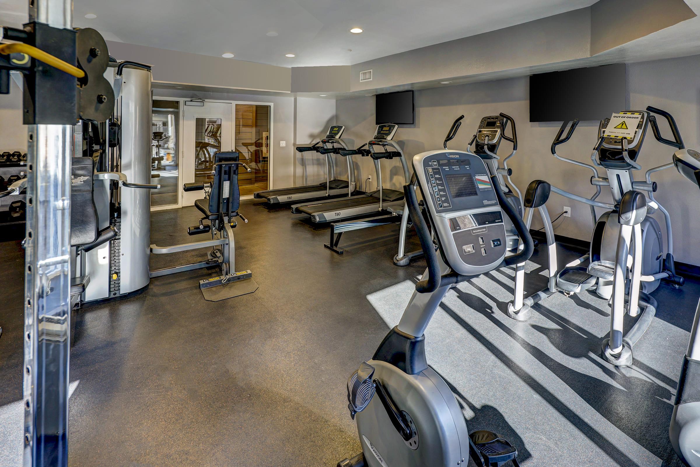 A modern gym interior featuring various exercise equipment, including treadmills, stationary bikes, weight machines, and an elliptical. The space is well-lit with large windows and has a clean, open layout. Two television screens are mounted on the walls for entertainment while working out.