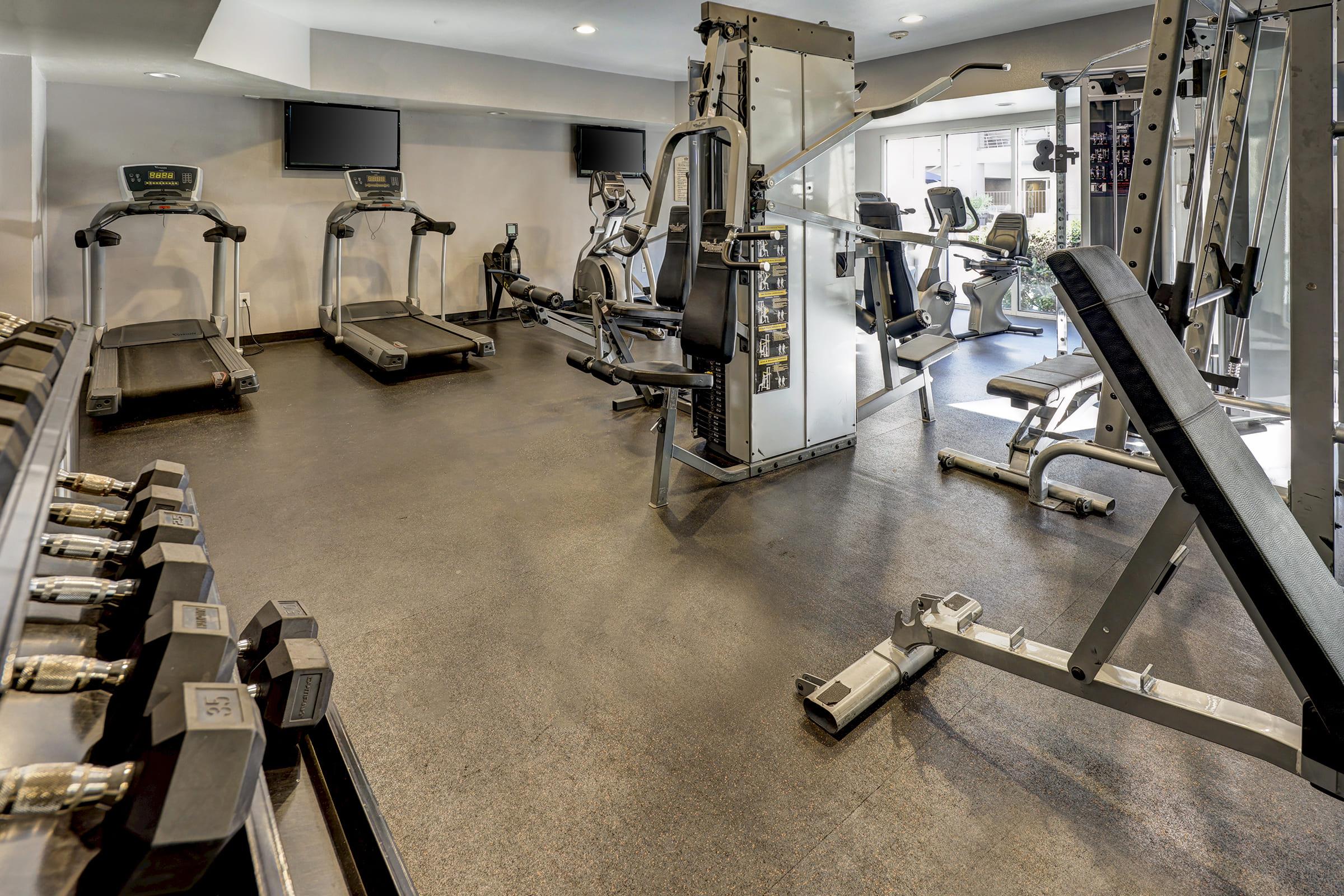 A modern gym interior featuring several cardio machines, including treadmills and exercise bikes, along with a weightlifting area that has free weights. The space is well-lit with large windows and is designed for fitness activities.