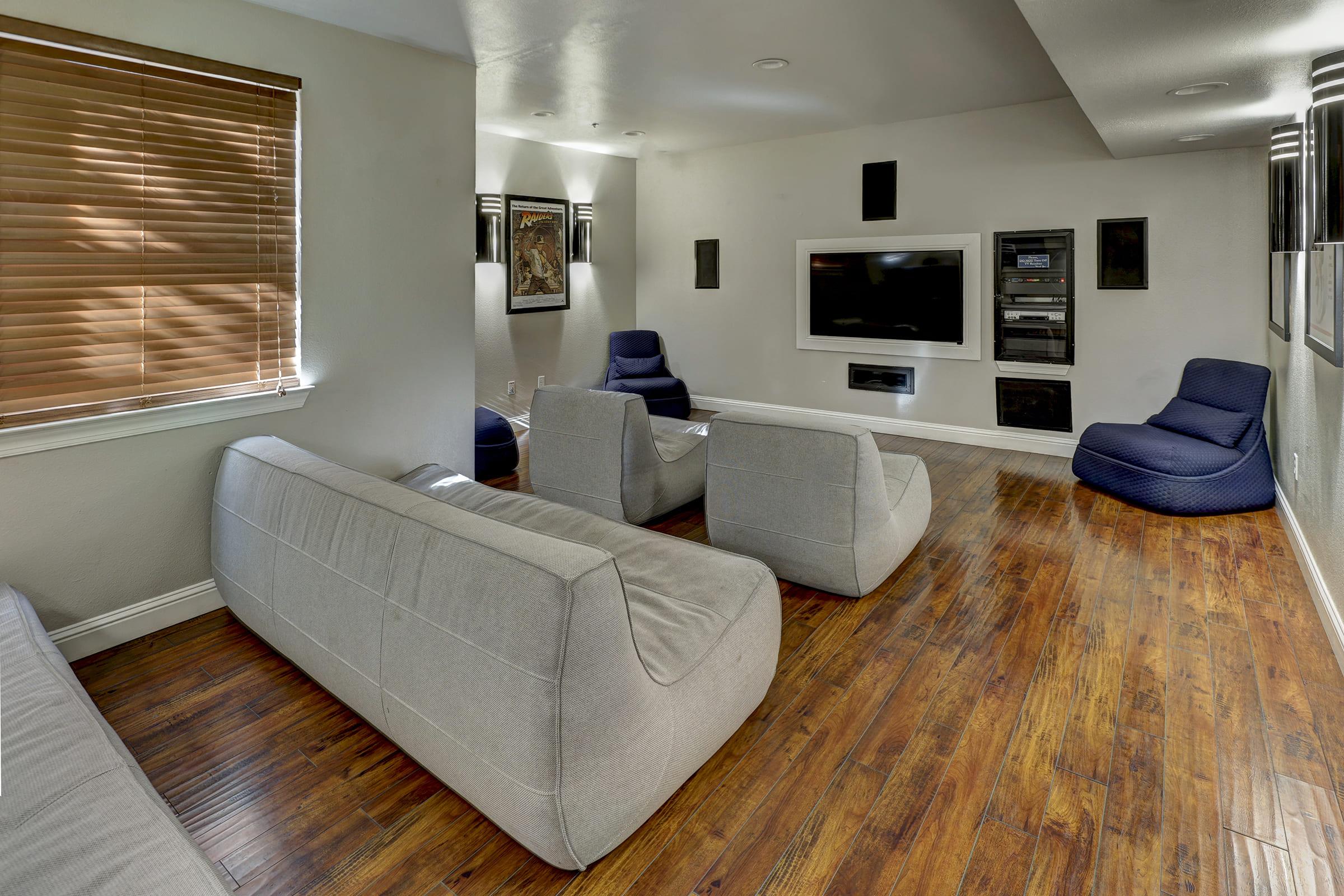 A modern living room featuring light gray sofas arranged facing a wall with a flat-screen TV and entertainment system. Soft blue bean bags are placed on the floor. The room has wooden flooring and natural light coming through a window with wooden blinds. Minimalist decor creates a cozy atmosphere.