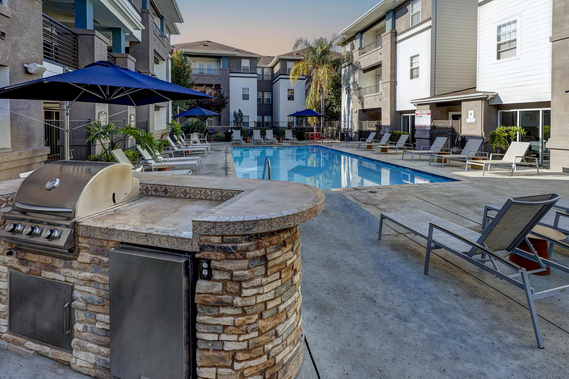 A serene outdoor pool area at an apartment community, featuring a large swimming pool surrounded by lounge chairs, shaded umbrellas, and a stone grill station. Lush greenery and modern architecture create a relaxing environment, ideal for leisure and social gatherings.