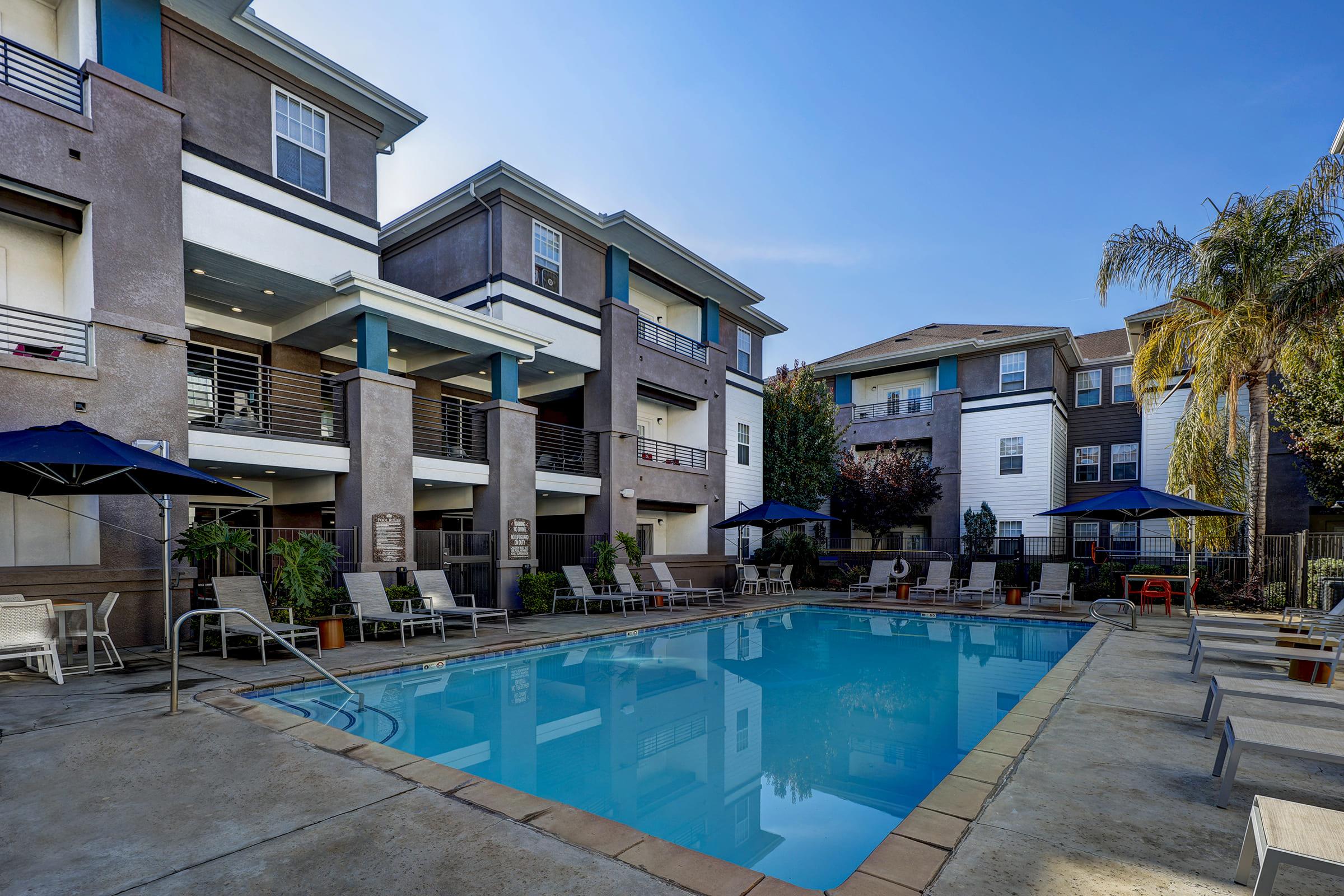 A sunny outdoor swimming pool surrounded by lounge chairs and shaded by umbrellas, with several multi-story apartment buildings in the background. Lush greenery and clear blue skies add to the inviting atmosphere of the pool area.