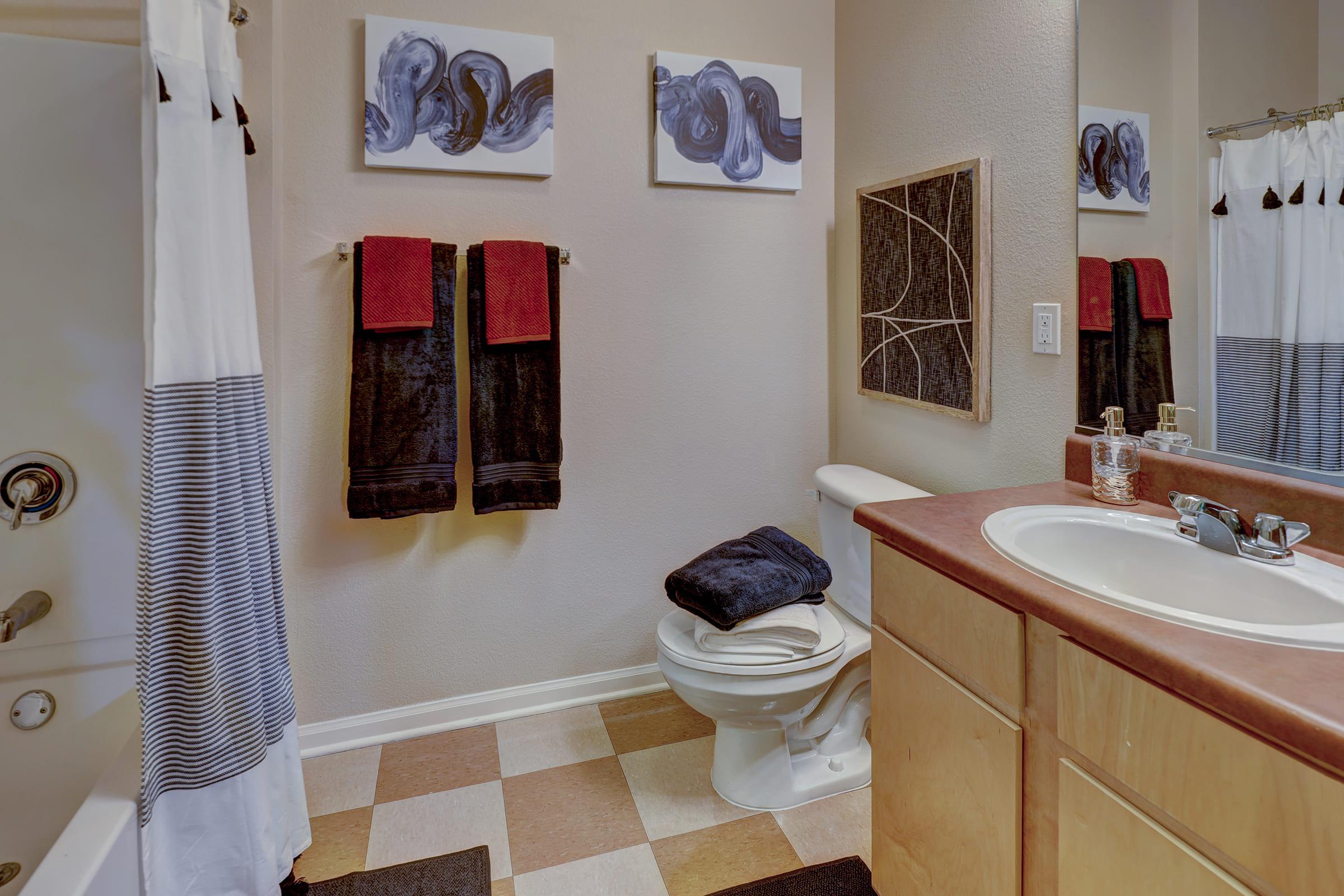 A modern bathroom featuring a toilet, sink, and mirror. The countertops are a warm brown, with a dark cabinet below. Decor includes abstract wall art, two hanging red and black towels, and a black rug on the floor. The shower is visible in the background, with a white curtain and striped pattern.