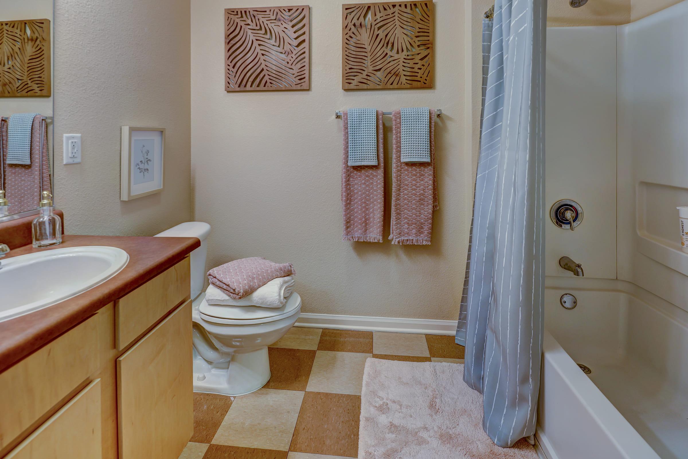 A cozy bathroom featuring a white toilet, a sink with wooden cabinets, and a bathtub with a shower curtain. The walls are adorned with decorative leaf-patterned art. Two towels hang on a rack, and soft towels are placed on the counter, adding a warm and inviting atmosphere. The floor has a checkered pattern in neutral tones.