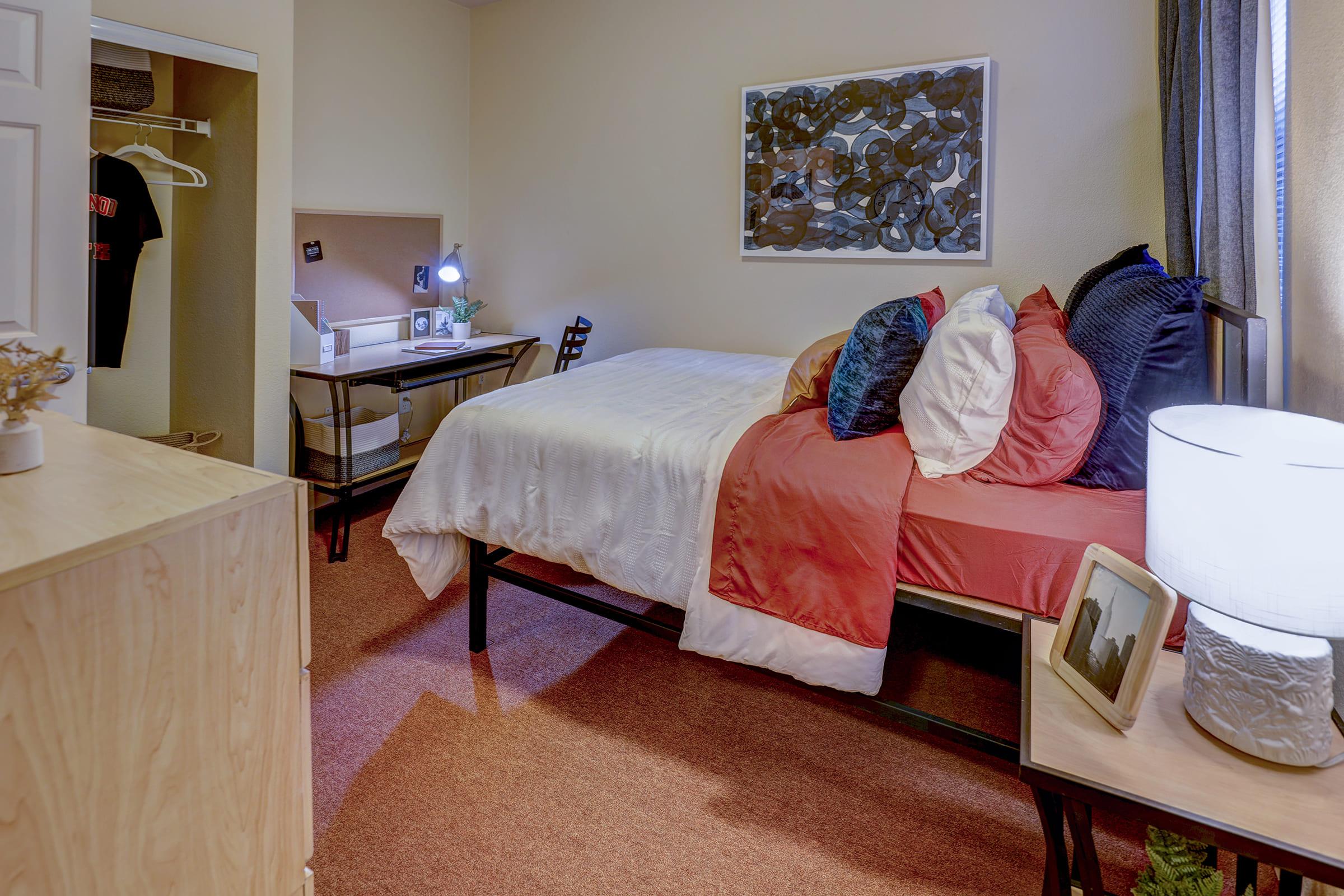 A cozy bedroom with a bed featuring a white and coral spread, colorful pillows, and a bedside lamp. A small desk with a chair and a few personal items is visible, along with a closet in the background. The room has warm lighting and neutral-colored walls, creating a welcoming atmosphere.