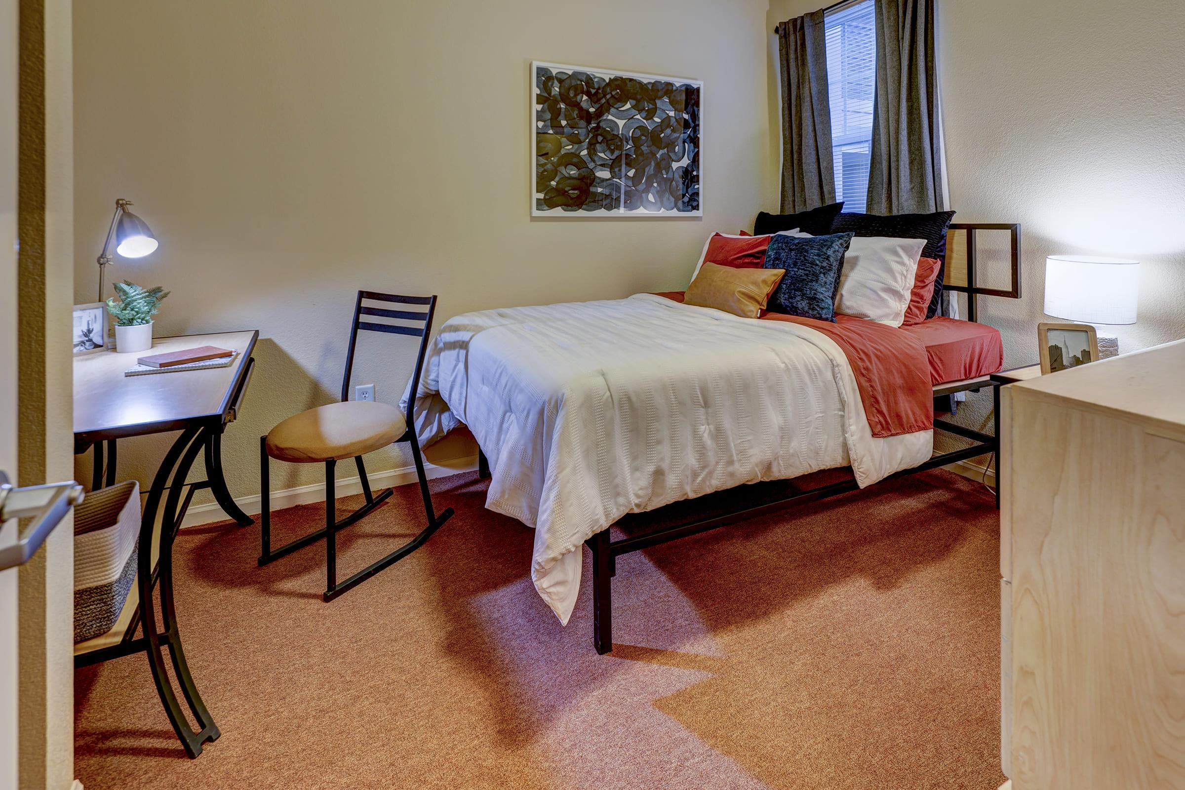 A cozy bedroom featuring a bed with red and white bedding, a nightstand with a lamp, a desk with a chair, and a window with curtains. The walls are a light color, and there's a piece of artwork above the bed, creating a warm and inviting atmosphere.