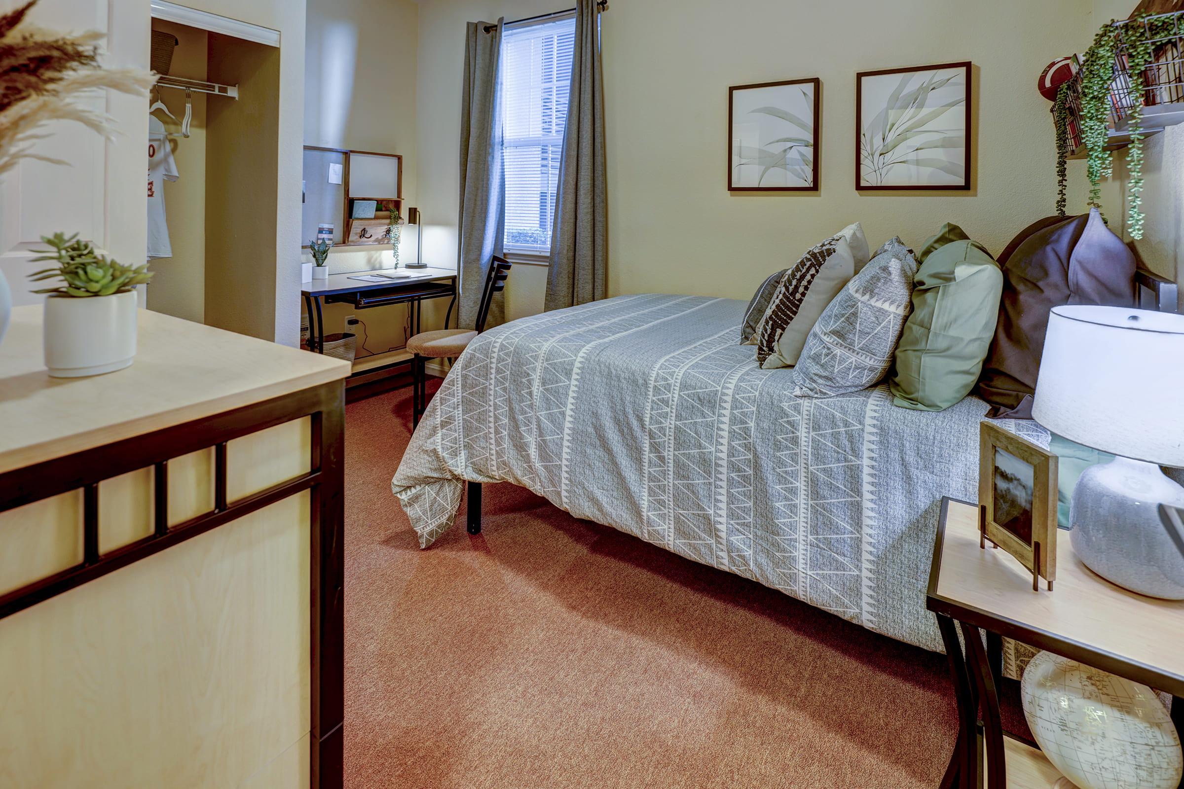 A cozy bedroom featuring a bed with patterned bedding, multiple decorative pillows, and bedside tables. A desk with a lamp is positioned near a window, and plants adorn the room. A closet is visible in the background, creating a warm and inviting atmosphere.