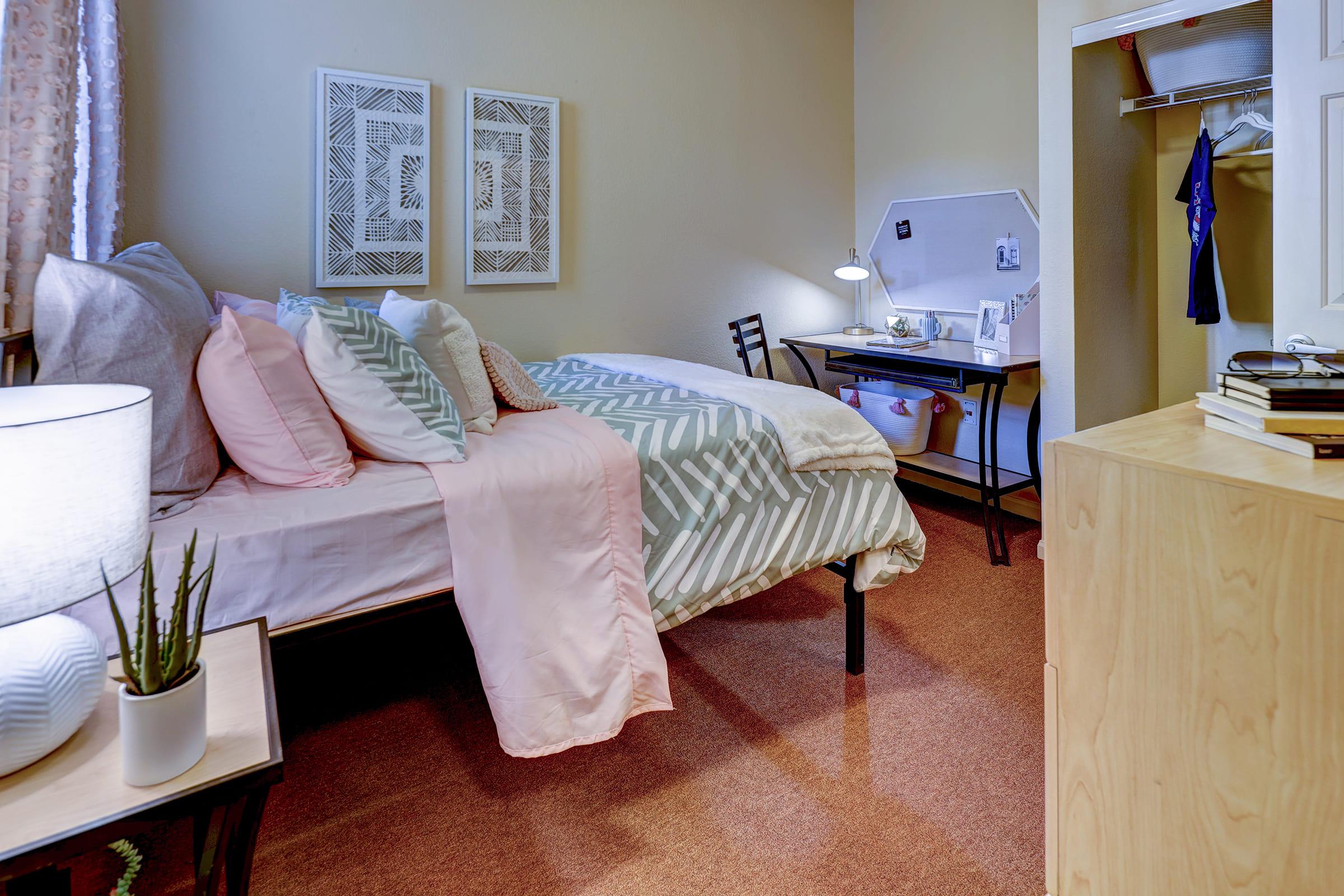 A cozy bedroom featuring a bed with patterned pillows and a pastel throw, a desk with personal items, a closet with clothes visible, and a bedside table with a lamp and a small plant. Soft lighting enhances the inviting atmosphere of the space.