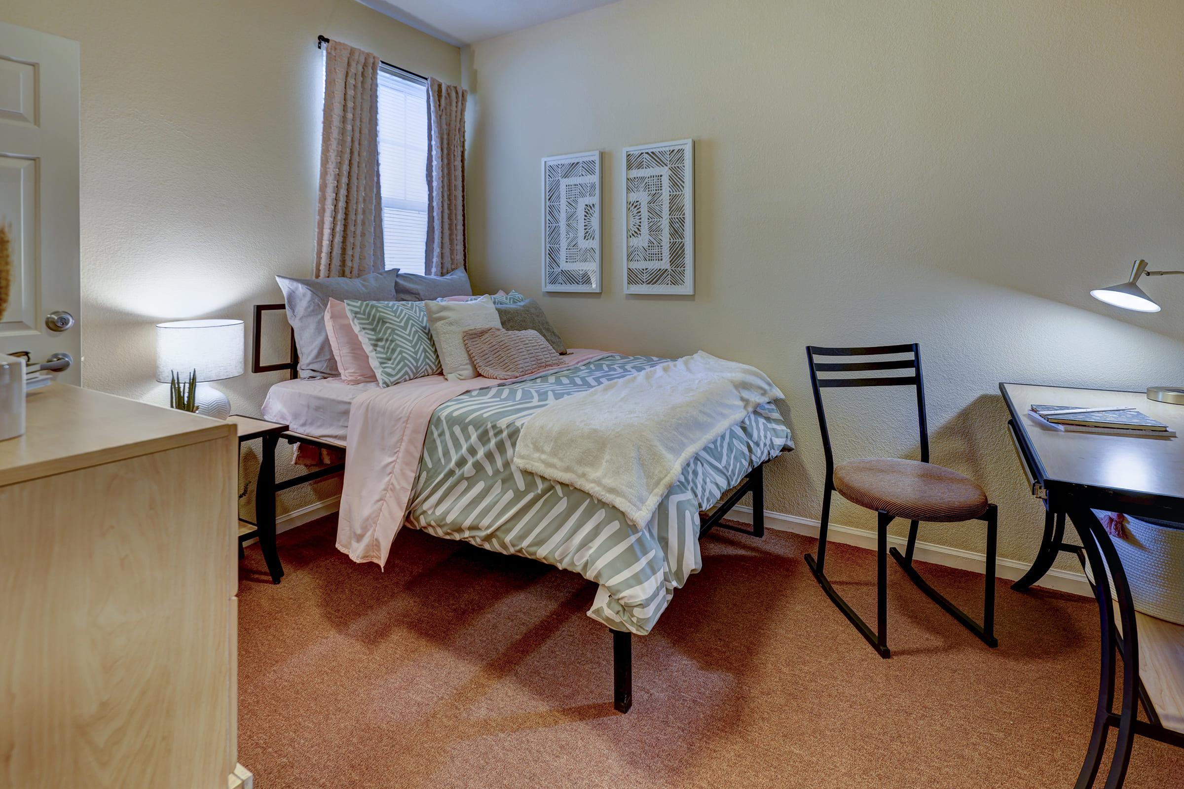 A cozy bedroom featuring a twin-sized bed with a mix of patterned pillows and a soft blanket. There is a small desk with a chair and a desk lamp, alongside a dresser. Light curtains frame the window, and two framed wall art pieces add a decorative touch. The room has neutral walls and a warm carpet.