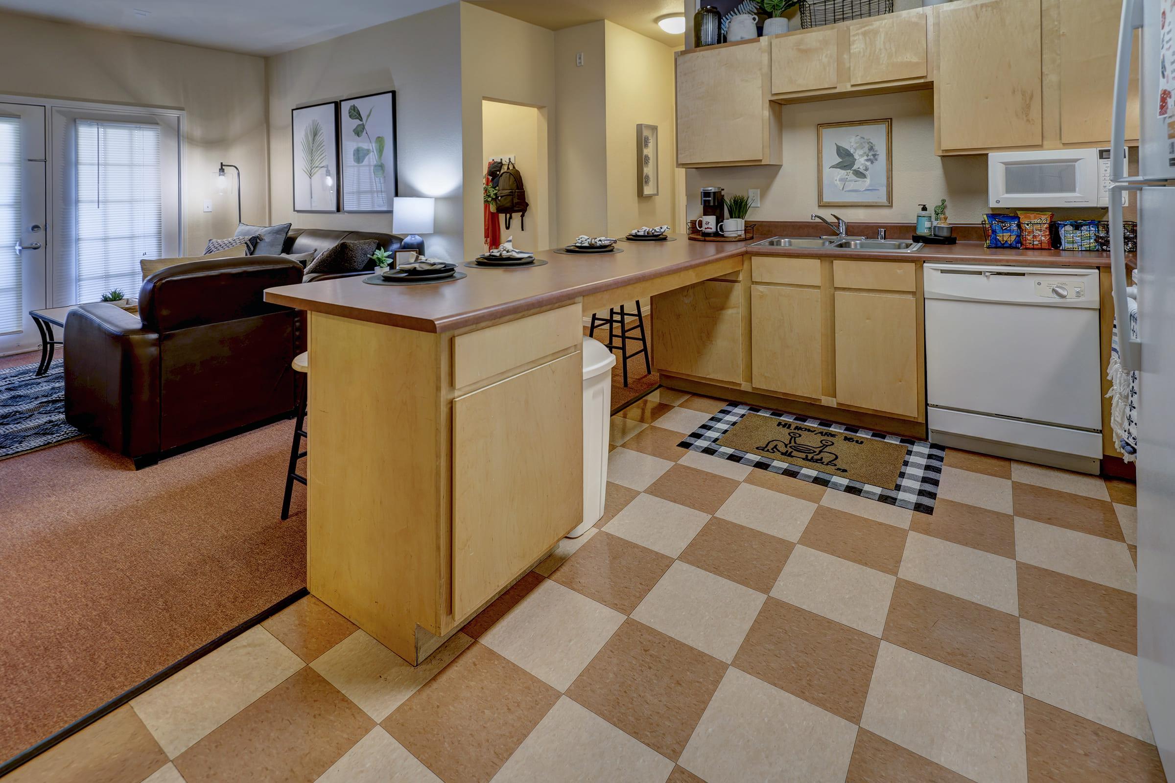 A cozy kitchen and living area with light wood cabinetry, a countertop with snacks, and a dishwasher. The kitchen features beige and brown checkered floor tiles. In the background, a comfortable living room is visible, furnished with a brown sofa and artwork on the walls, along with sliding glass doors leading outside.