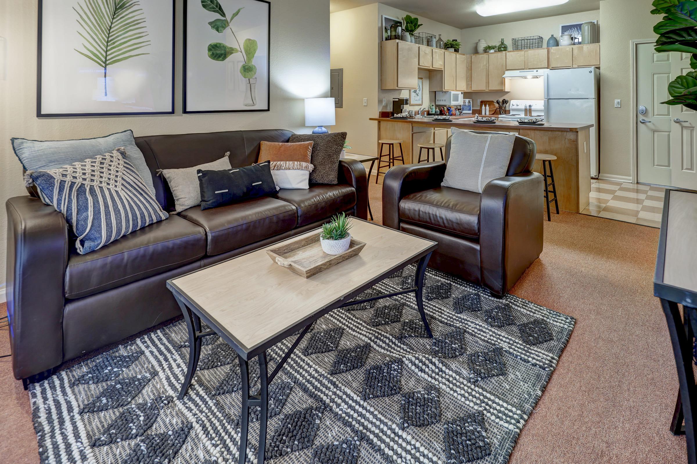 A cozy living room featuring a brown leather sofa with decorative pillows, a wooden coffee table atop a patterned rug, and a comfortable armchair. In the background, a kitchen with wooden cabinetry and modern appliances is visible, along with green plants and wall art depicting leaves.