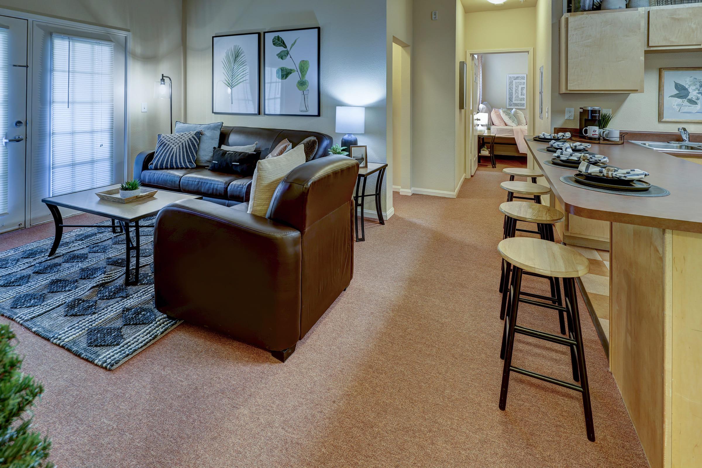 A cozy living area featuring a dark leather sofa, a coffee table with a decorative plant, and a patterned rug. There are two bar stools at a kitchen counter, with diningware set out. The space is warmly lit, with wall art and a glimpse of a bedroom in the background.