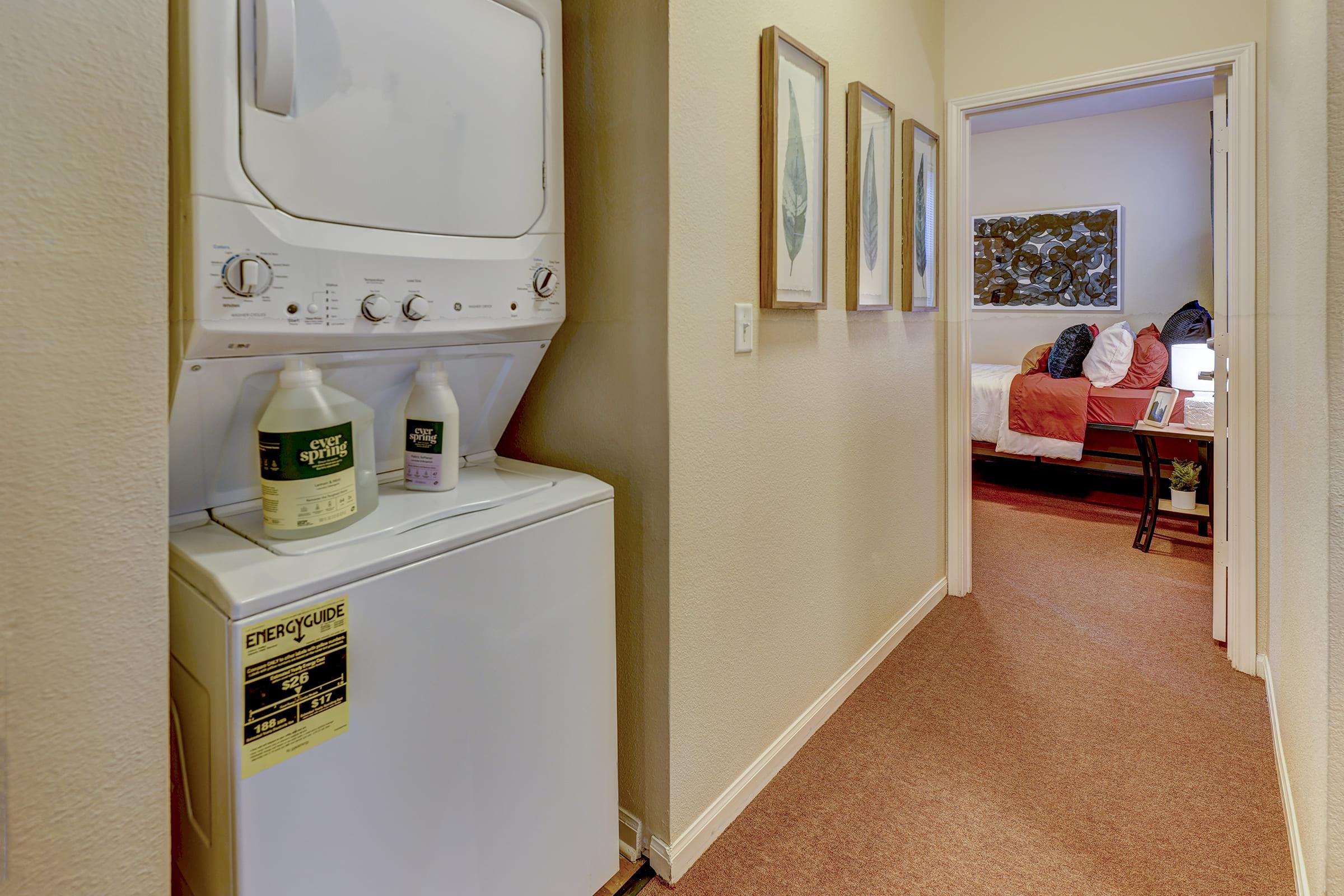 A small hallway featuring a stacked washer and dryer unit on the left. Two laundry detergent bottles are placed on top of the dryer. In the background, a bedroom is partially visible, complete with a bed and decorative wall art. The walls are painted beige, and the carpet is a warm color.