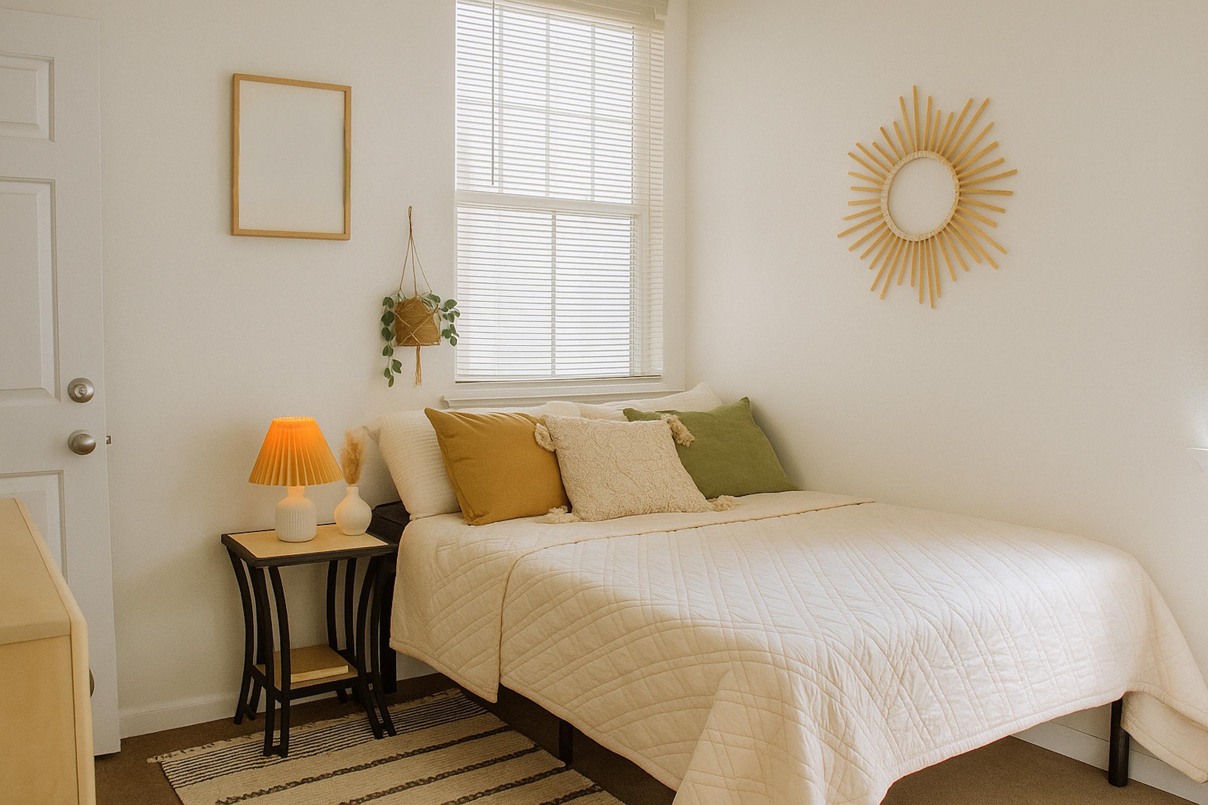 A cozy bedroom featuring a queen-sized bed with a white quilt, adorned with green and beige pillows. A small side table holds a lamp, and a wall mirror is framed in sunburst design. Natural light filters through the window, complemented by simple decor and a few plants.