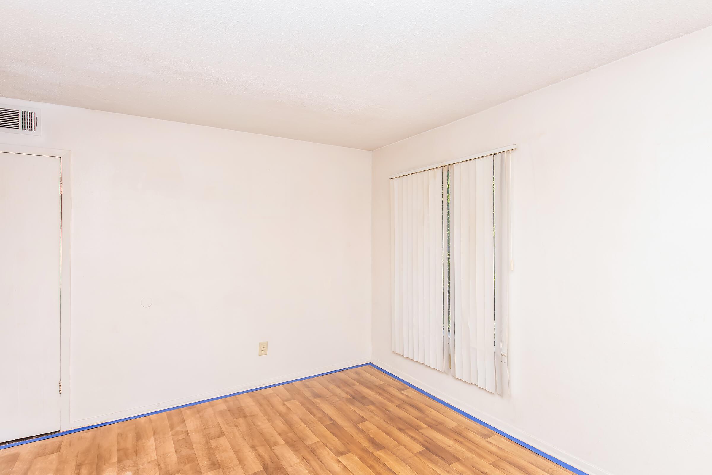 A simple, empty room with light-colored walls and a wooden floor. There is a closed door on one side and a window with vertical blinds allowing natural light to enter. The floor has blue painter's tape along the edges, indicating preparation for painting or renovations.