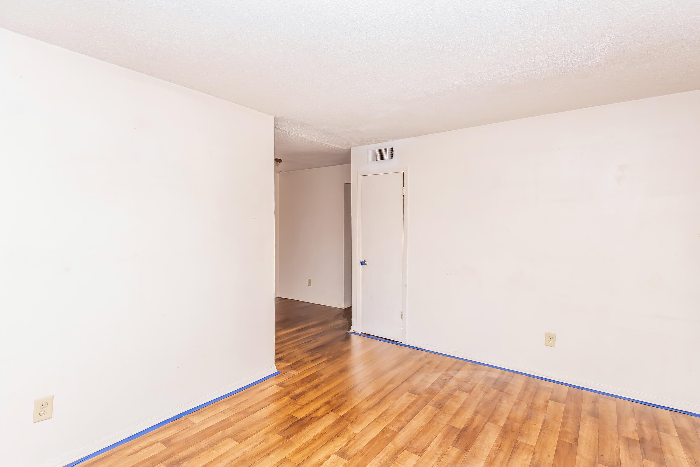 An empty room with light-colored walls and a wooden floor. The space features a door on one side and a smooth, unadorned wall opposite. Blue painter's tape outlines the floor along the wall, indicating preparation for painting or renovation. The lighting is bright and the atmosphere is clean and open.