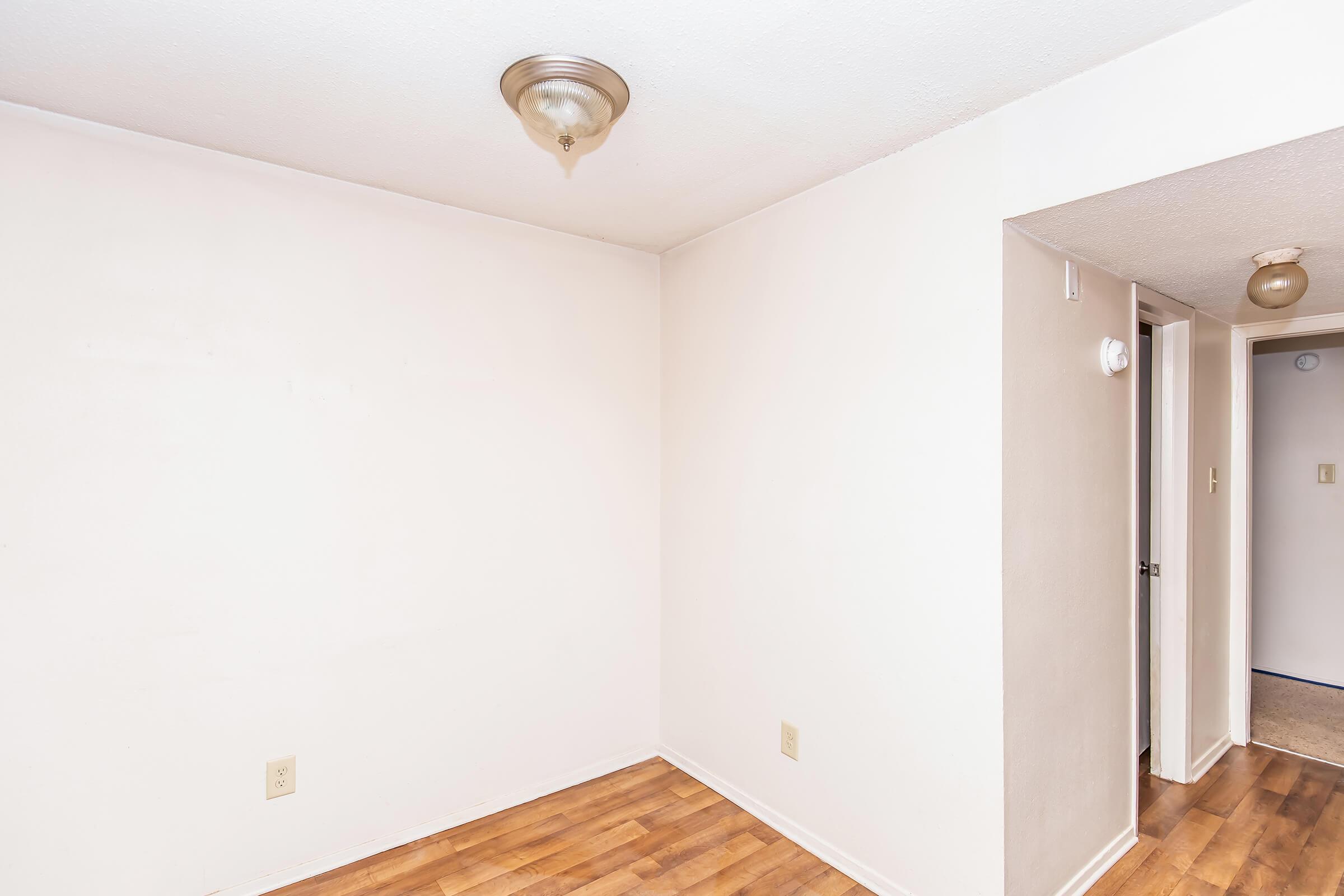 A vacant room with light-colored walls and hardwood flooring. There is a ceiling light fixture in the center, and a doorway leading to another room is visible on the right side. The overall ambiance is bright and minimalistic, with no furniture present.