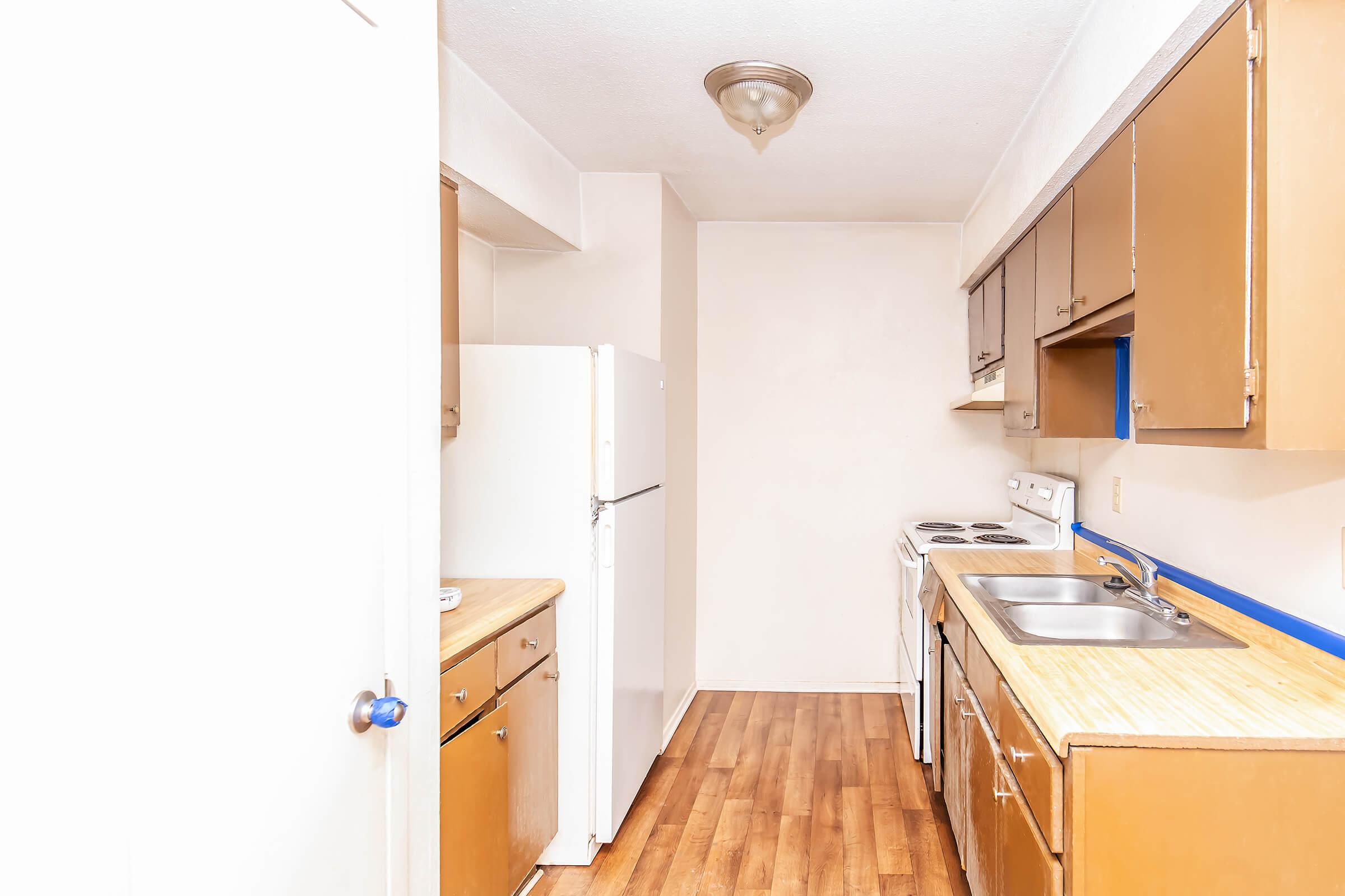 A small kitchen featuring light brown wooden cabinets, a white refrigerator, a white stove, and a double sink. The floor is made of light wood, and the walls are mostly white, with blue accents near the sink area. The space is well-lit, suggesting a cozy and functional design.