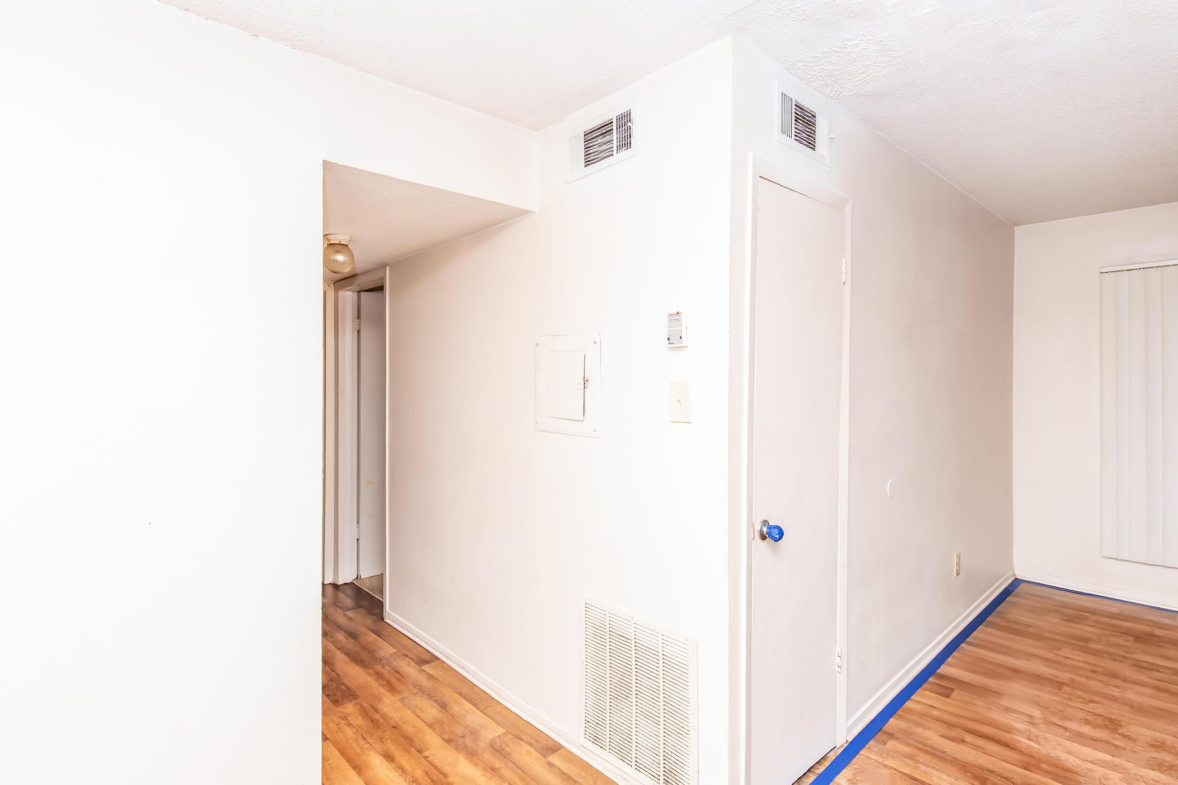 A well-lit, minimally furnished interior space featuring a white wall with a blue door knob, a vent near the floor, and a textured ceiling. To the left, there is an open door leading to another room, and the floor is covered with light brown laminate flooring. The walls are painted white, giving a clean and bright appearance.