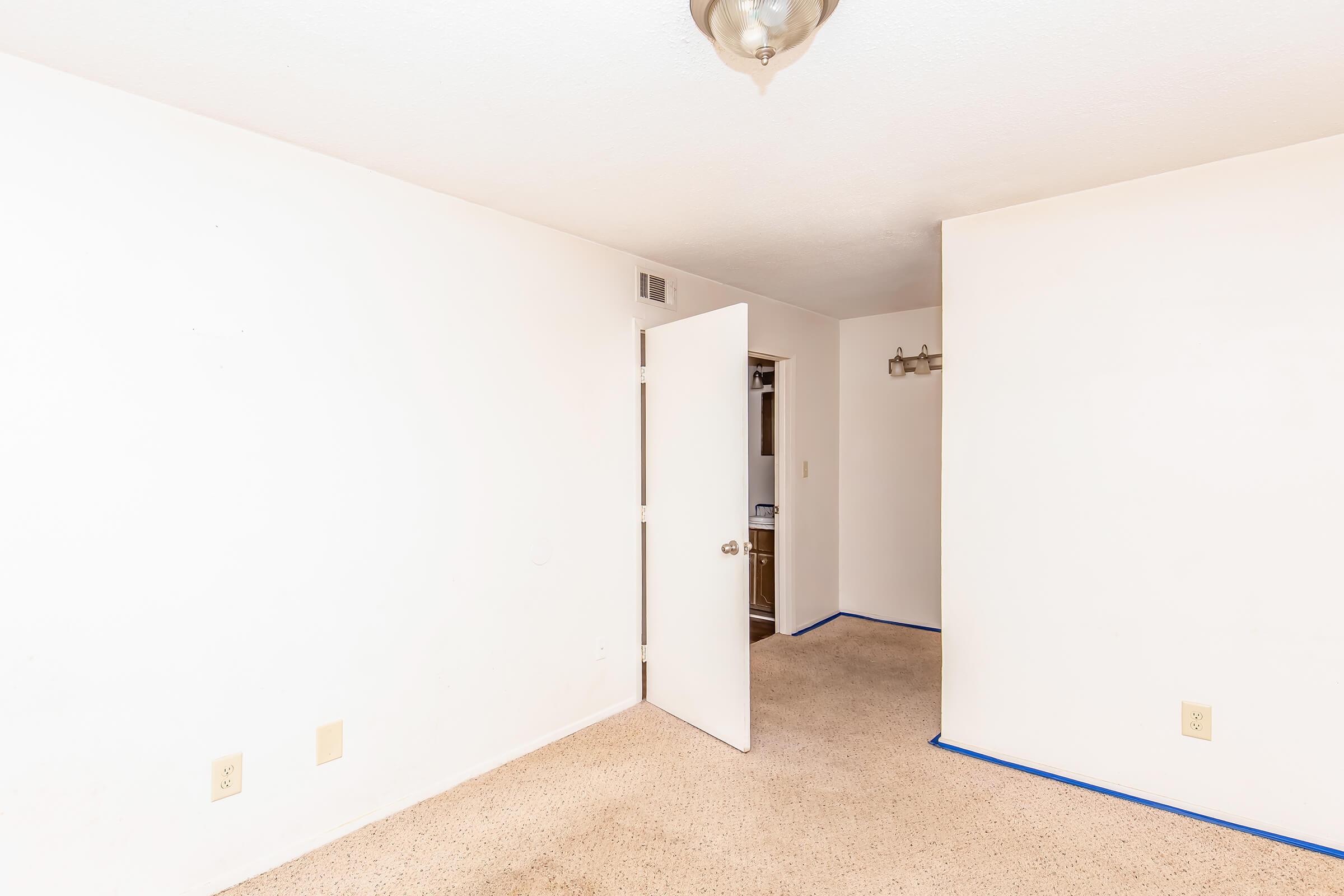 A vacant, empty room with light-colored walls and a carpeted floor. There is a ceiling light fixture and an open door leading to a closet. The room appears clean and well-lit, with no furniture or decorations present.