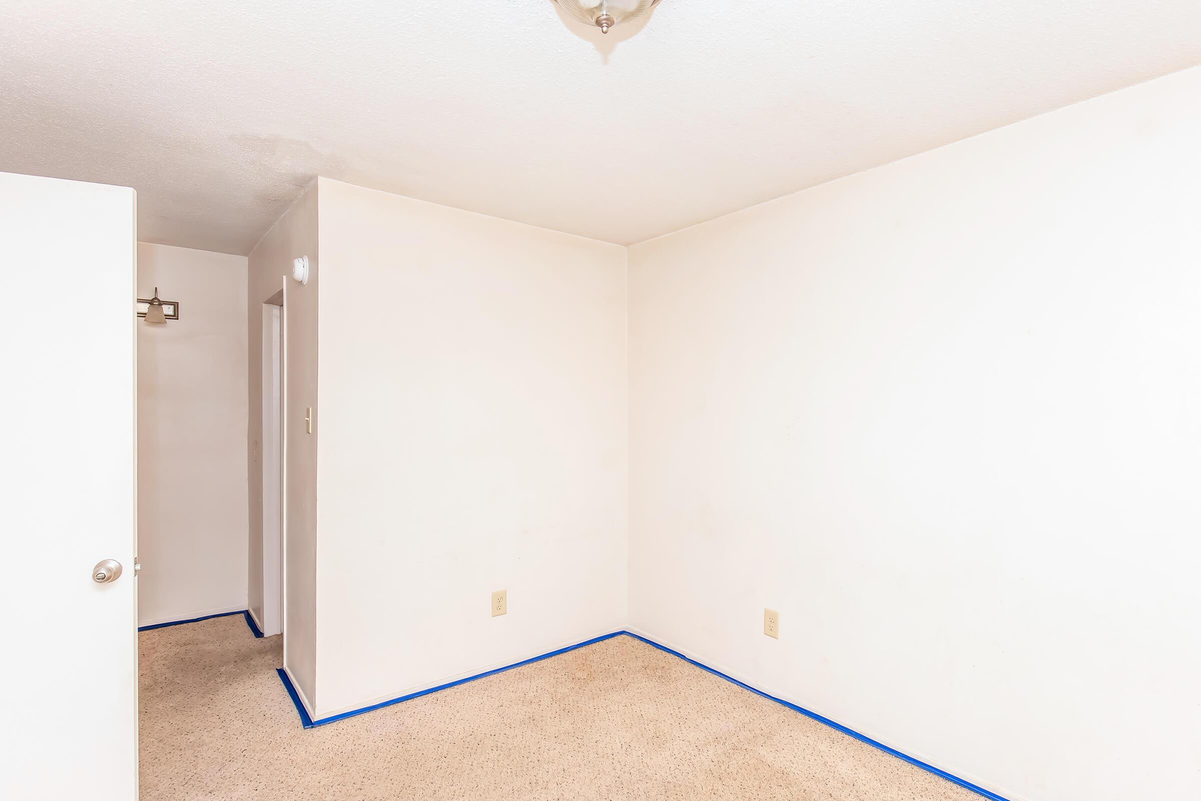 A small, empty room with beige carpet and white walls. There is a door to the left and a small hallway visible through another door on the right. Blue painter's tape lines the base of the walls, indicating preparation for painting. The ceiling has a simple light fixture.