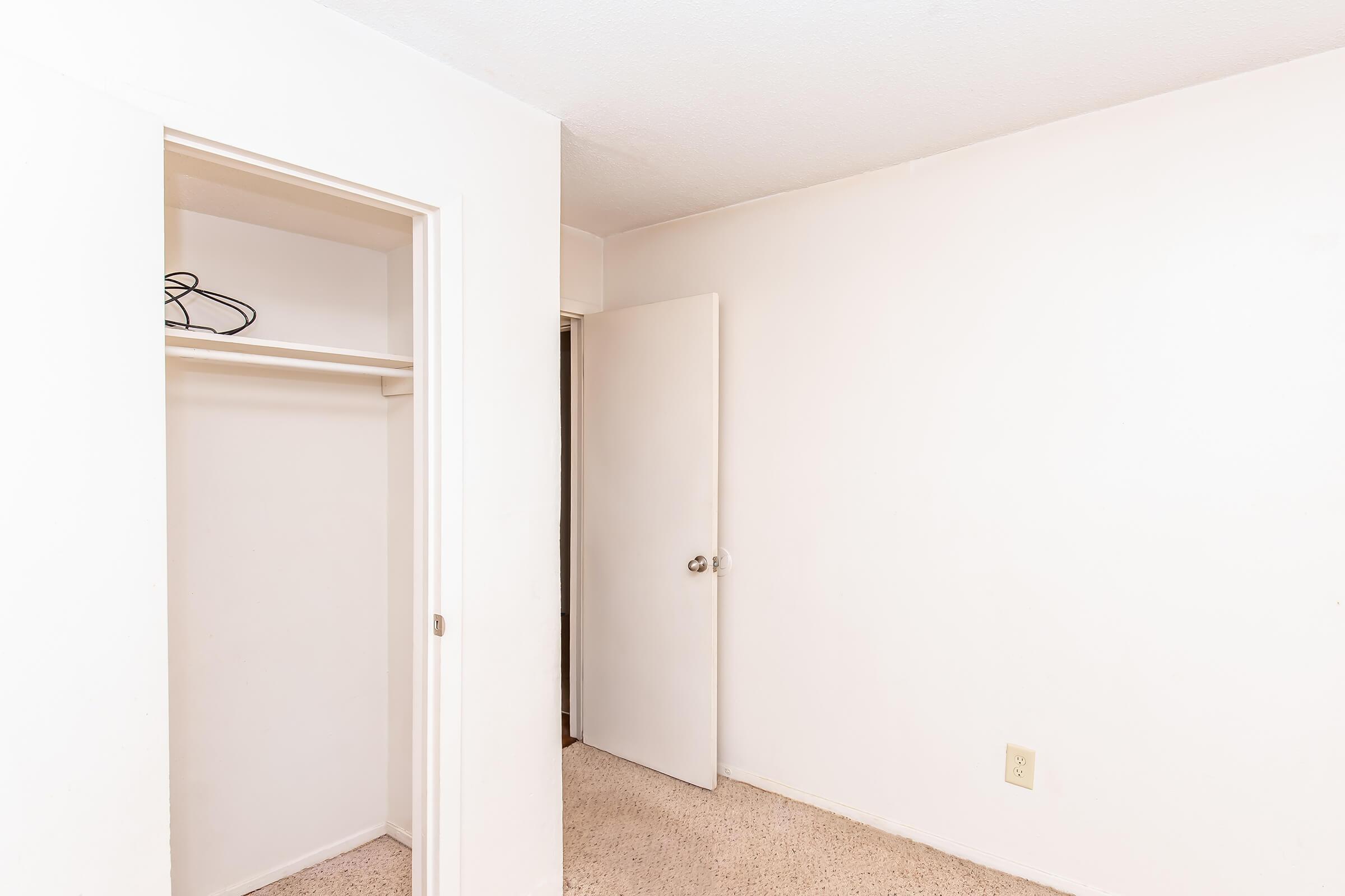 A small, empty room with light beige walls and carpet. There is a closet with a hanger inside, and a closed door leading to another space. The room has a clean and simple appearance, with no furniture or decorations.