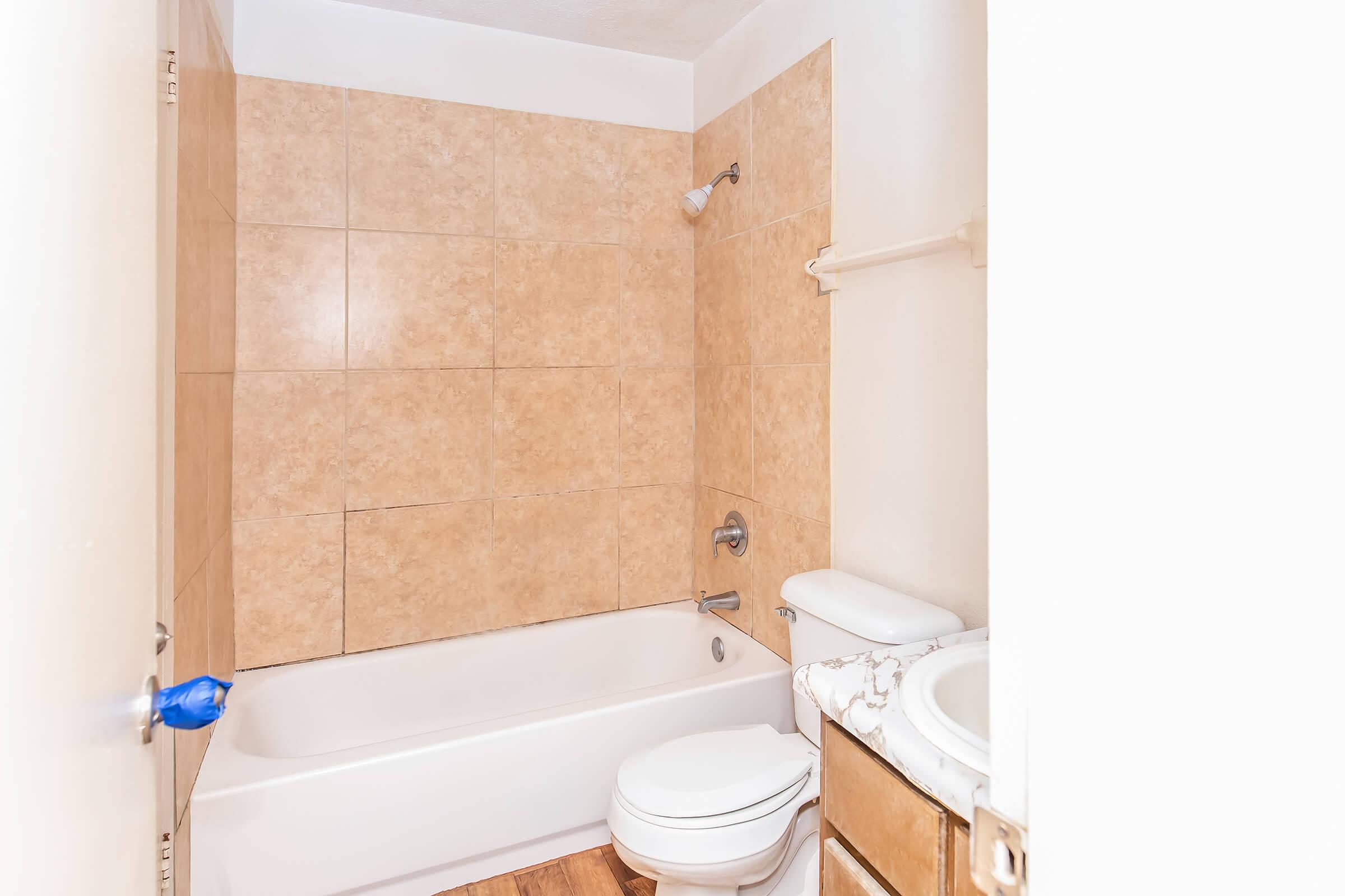 A small bathroom featuring beige tiled walls, a white bathtub, a toilet, and a sink. The bathtub is equipped with a showerhead, and there is a wooden floor. The overall design is simple and functional, with light colors creating a spacious feel.