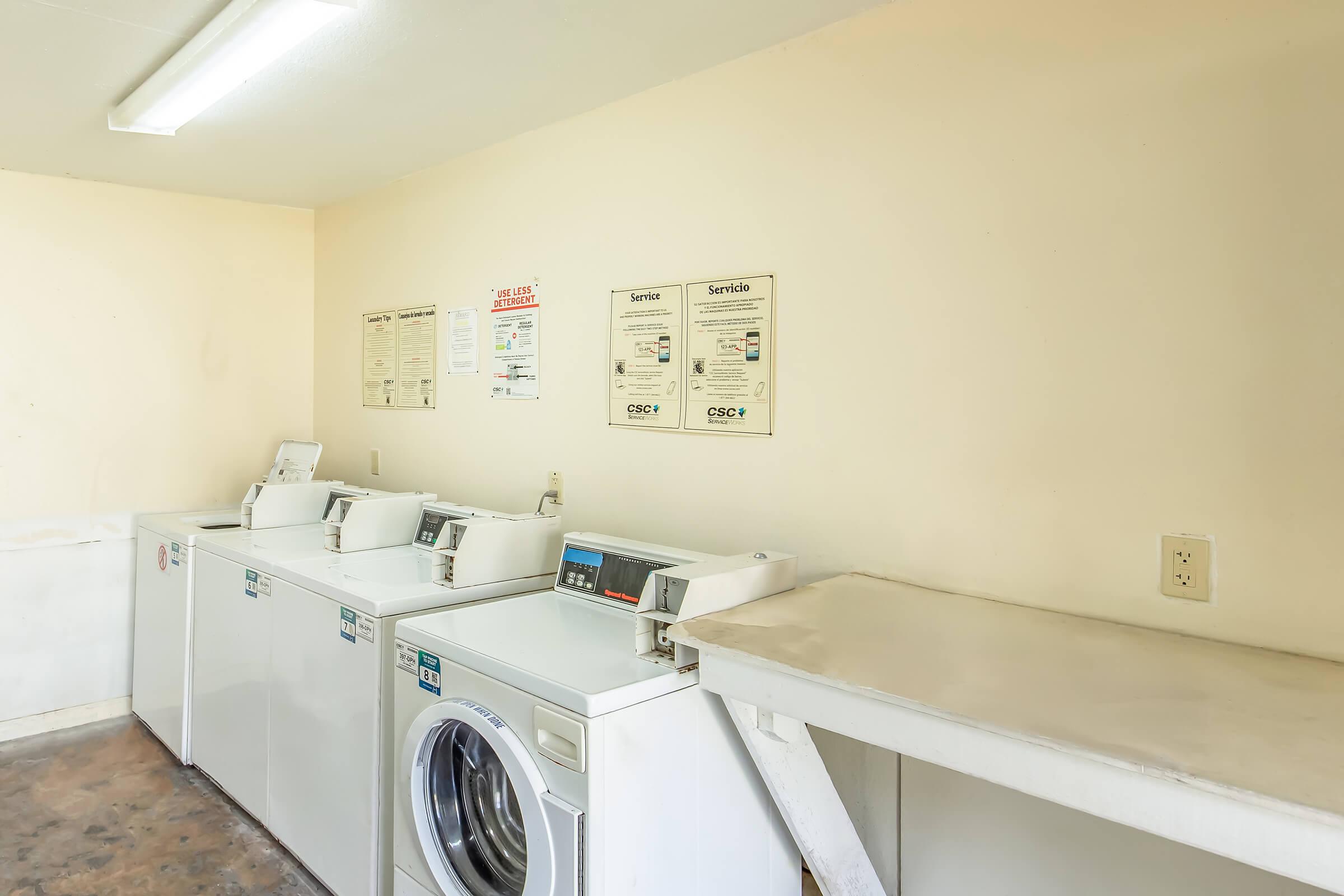 A clean and well-lit laundry room featuring several white washing machines and a dryer. There is a folding table on the right side, and informational posters are mounted on the wall. The floor appears to be a simple, unfinished surface.