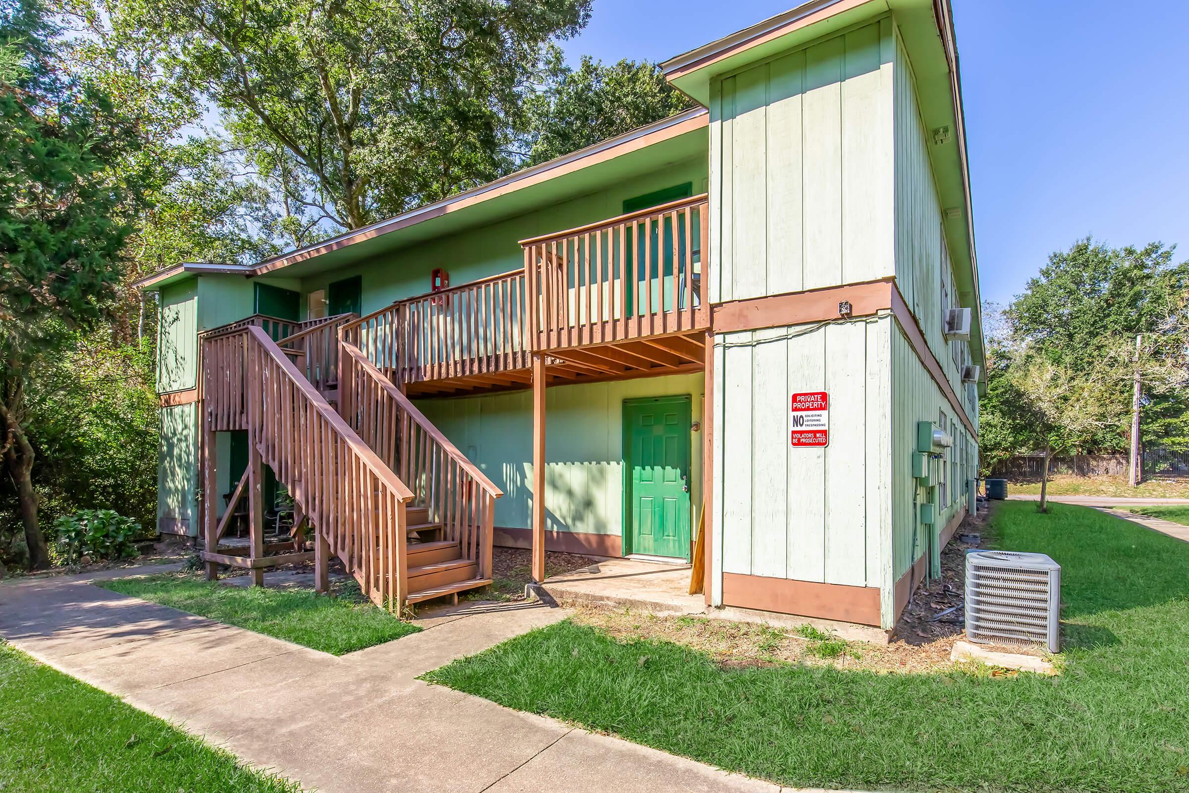 A two-story residential building with a light green exterior and brown trim. It has a wooden staircase leading to a second-floor entrance and a grassy area in front. Utility meters are visible on the side, and there is an air conditioning unit near the sidewalk. Trees are in the background, providing some shade.