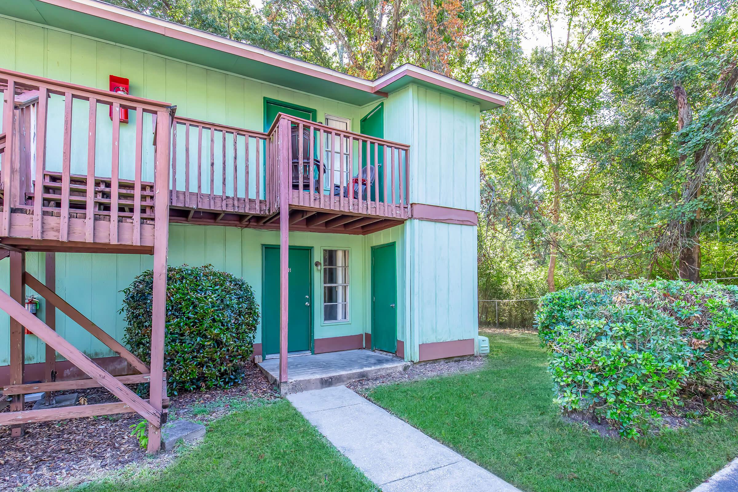 A light blue two-story building with green doors, featuring a wooden balcony and steps. The area is surrounded by lush greenery, including bushes and trees. A concrete pathway leads to the entrance, creating a welcoming outdoor space.