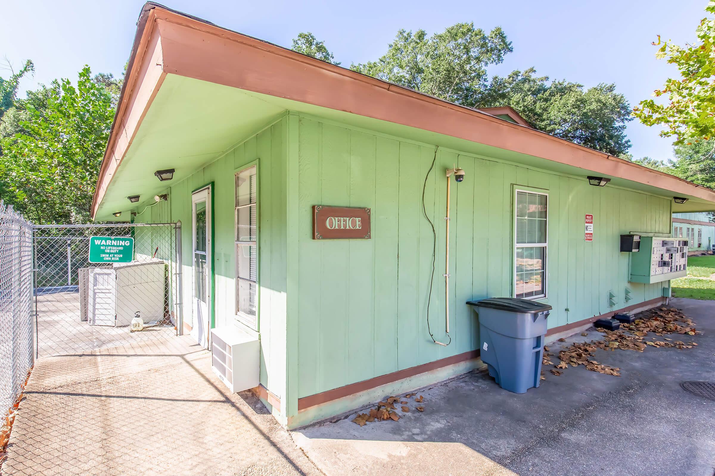 Small, single-story green office building with a sign labeled "OFFICE" on the wall. It has several windows and is surrounded by trees. A gray trash bin is placed beside the building, and there is a fenced area nearby with a warning sign visible. The ground is paved with light-colored stones.
