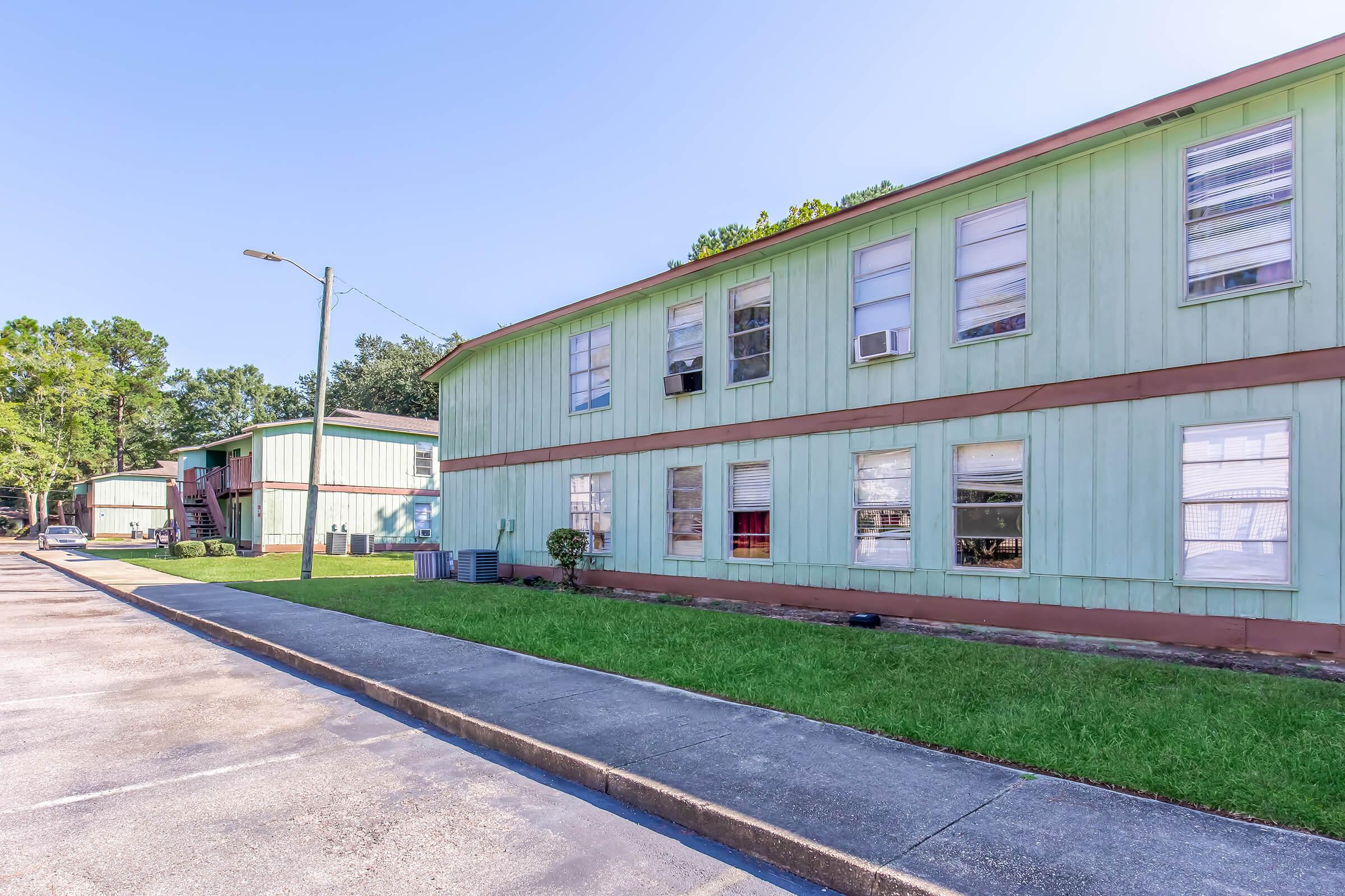 A green two-story apartment building with multiple windows, surrounded by a grassy area and trees. A sidewalk runs alongside the building, with a light post visible in the foreground. The setting appears to be in a residential neighborhood with clear blue skies.