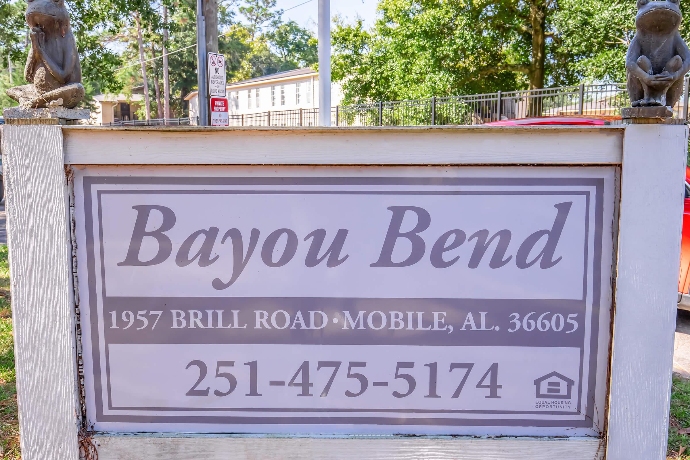 Sign for Bayou Bend, located at 1957 Brill Road, Mobile, AL 36605. The sign features contact information: 251-475-5174. Surrounding the sign are decorative elements, including two small sculptures. The background includes trees and a building.